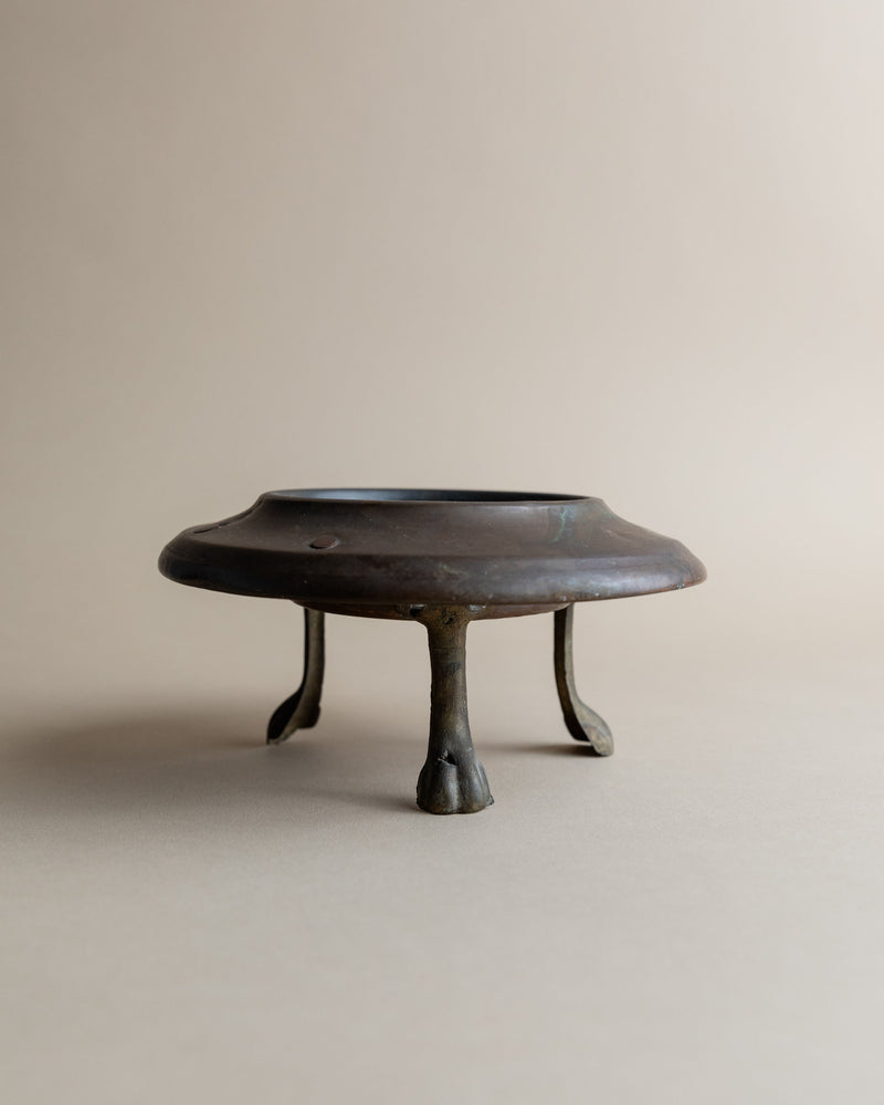 The Bottega Jacobs Vintage Copper Candle Holder, featuring a handcrafted design with a flat circular top and three hoof-like curved legs, is displayed on a neutral background, embodying brutalist design.