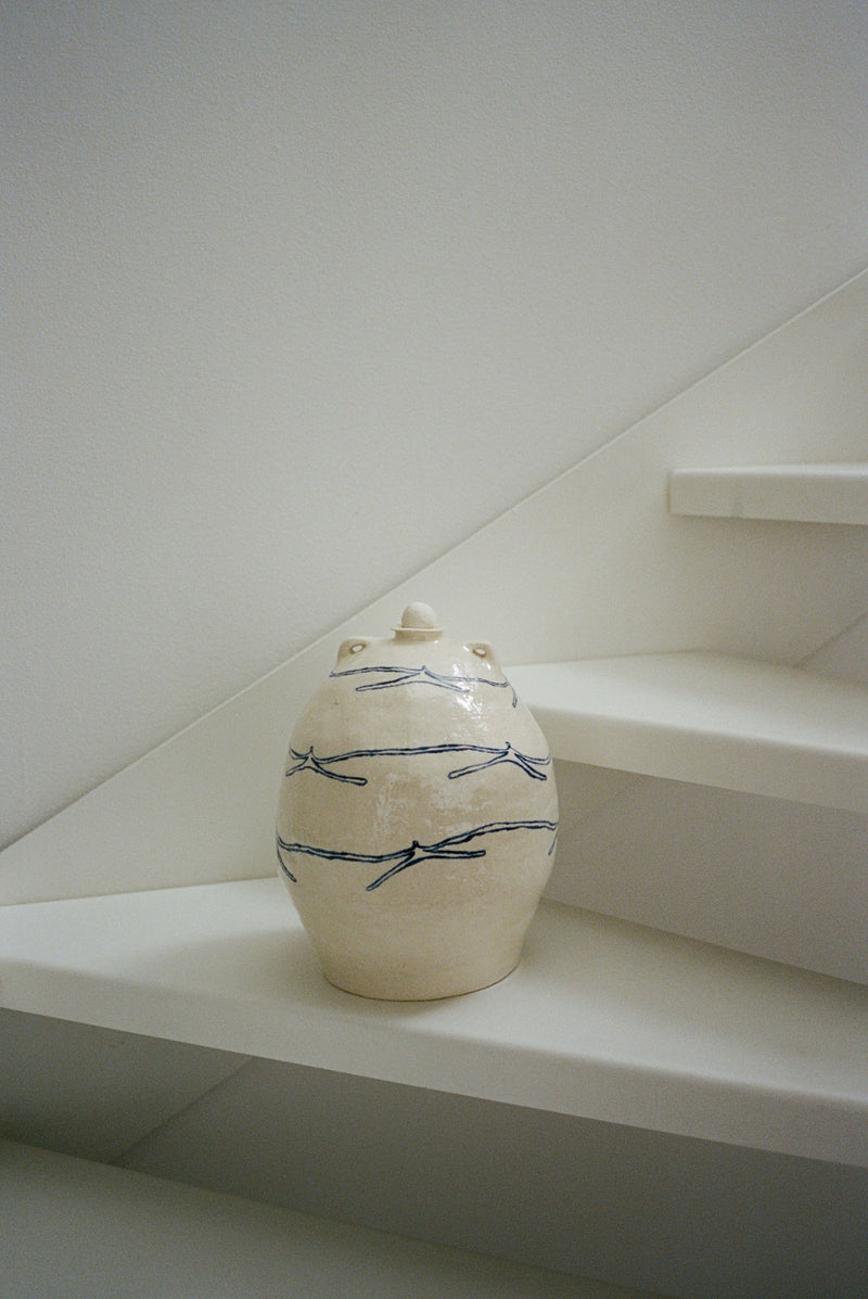 The 'Ladies Holding Hands' Ceramic Vase by ATELIER1991, cream-colored with simple blue dragonfly designs and a lid, sits on a white staircase against a plain wall.