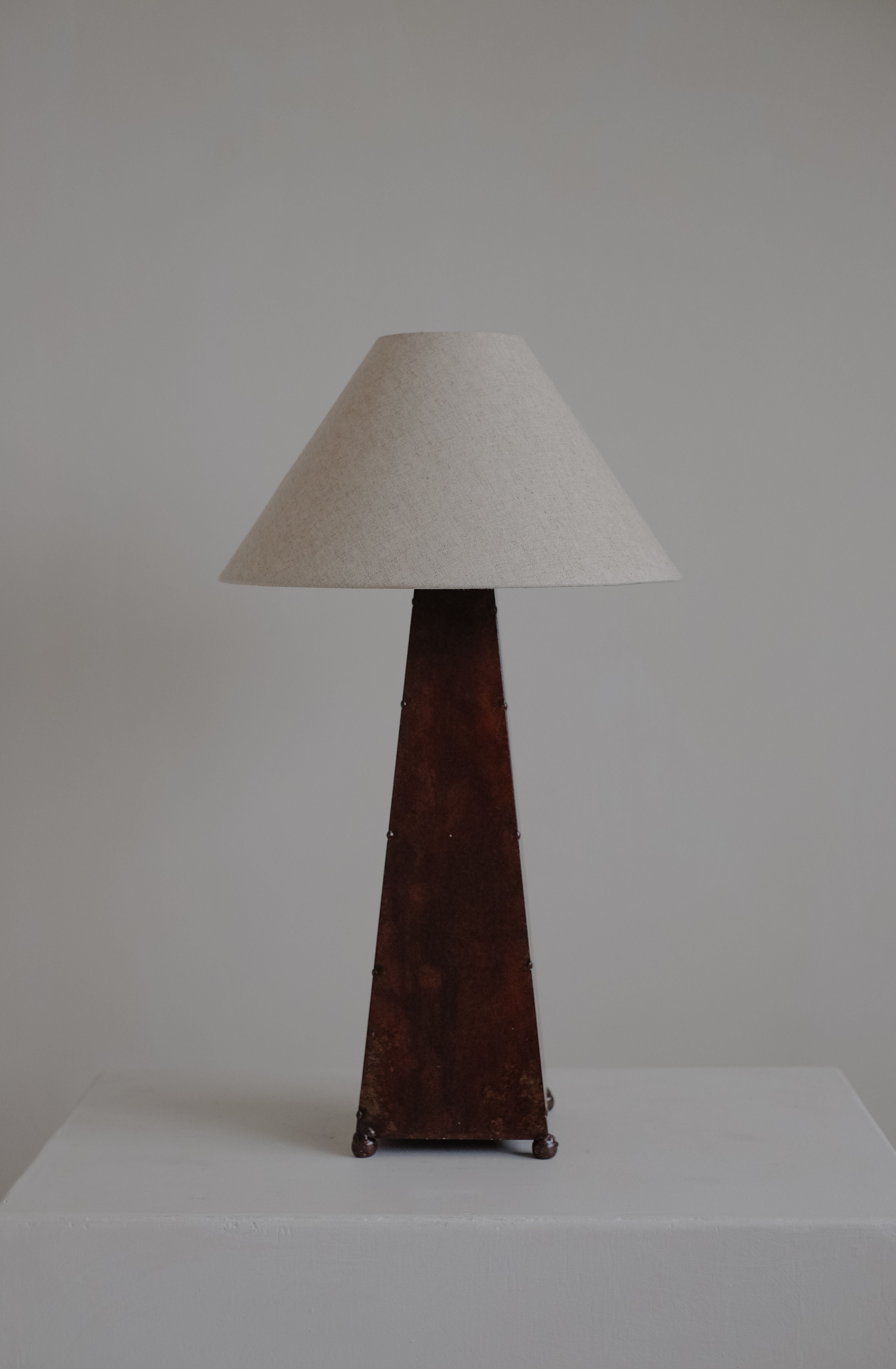 A tall, vintage septembre studios Obelisk Table Lamp with a pyramid-shaped wooden base featuring a subtle rust-copper patina and a wide, conical beige lampshade sits on a white surface against a light gray background.