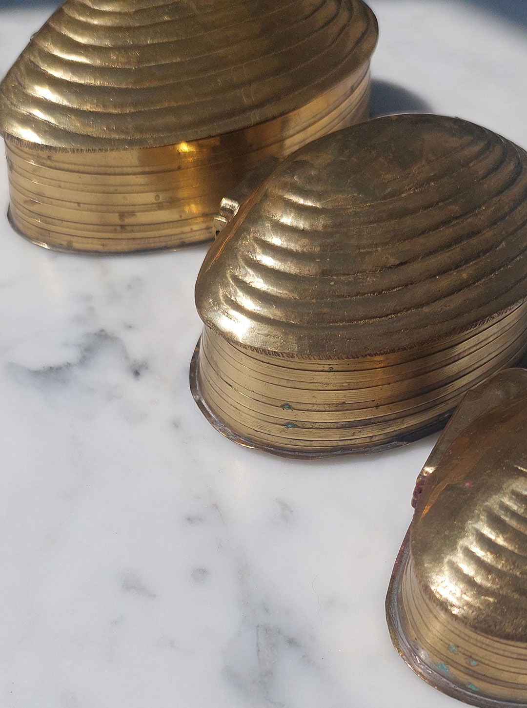 Les Objoies Set of 3 Nesting Shell Boxes with a brass gold finish are seen on a white marble surface under natural light. The boxes are varied in size, stacked slightly, and gleaming with a polished metallic finish. Shadows indicate the direction of the sunlight.