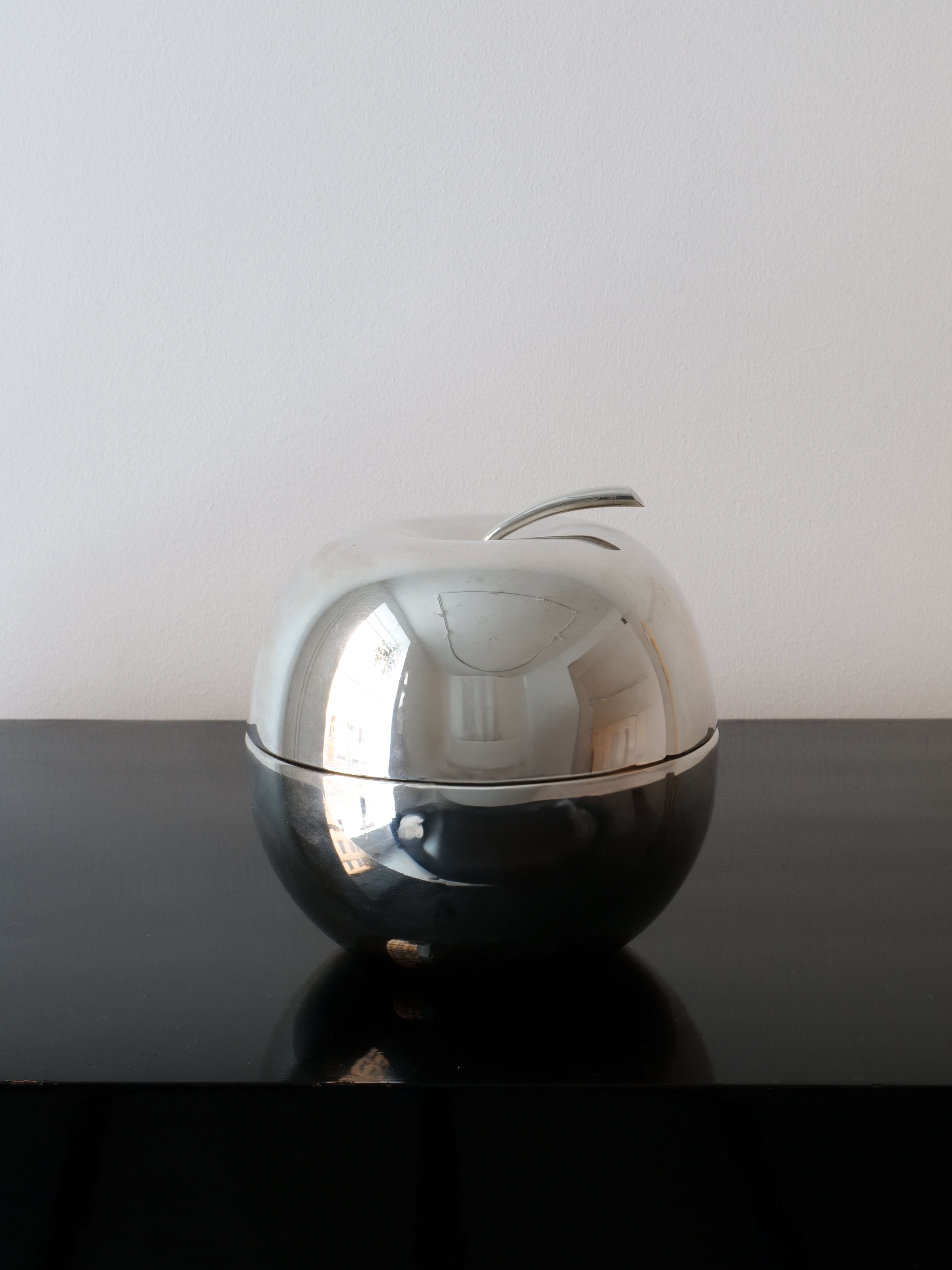 On a black surface against a plain white background rests the Christofle Apple Jar Lino Sabattini by Collection apart. This modernist apple-shaped container features a shiny, metallic exterior that beautifully reflects subtle light and room nuances, enhancing its elegant allure.