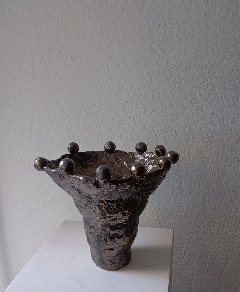 The Coupe Lilibeth 3 by Claire Pain is a unique metallic textured ceramic vase with a flared rim and rounded protrusions, displayed on a white pedestal against a light textured wall, evoking sculptural bronze enamel artistry.