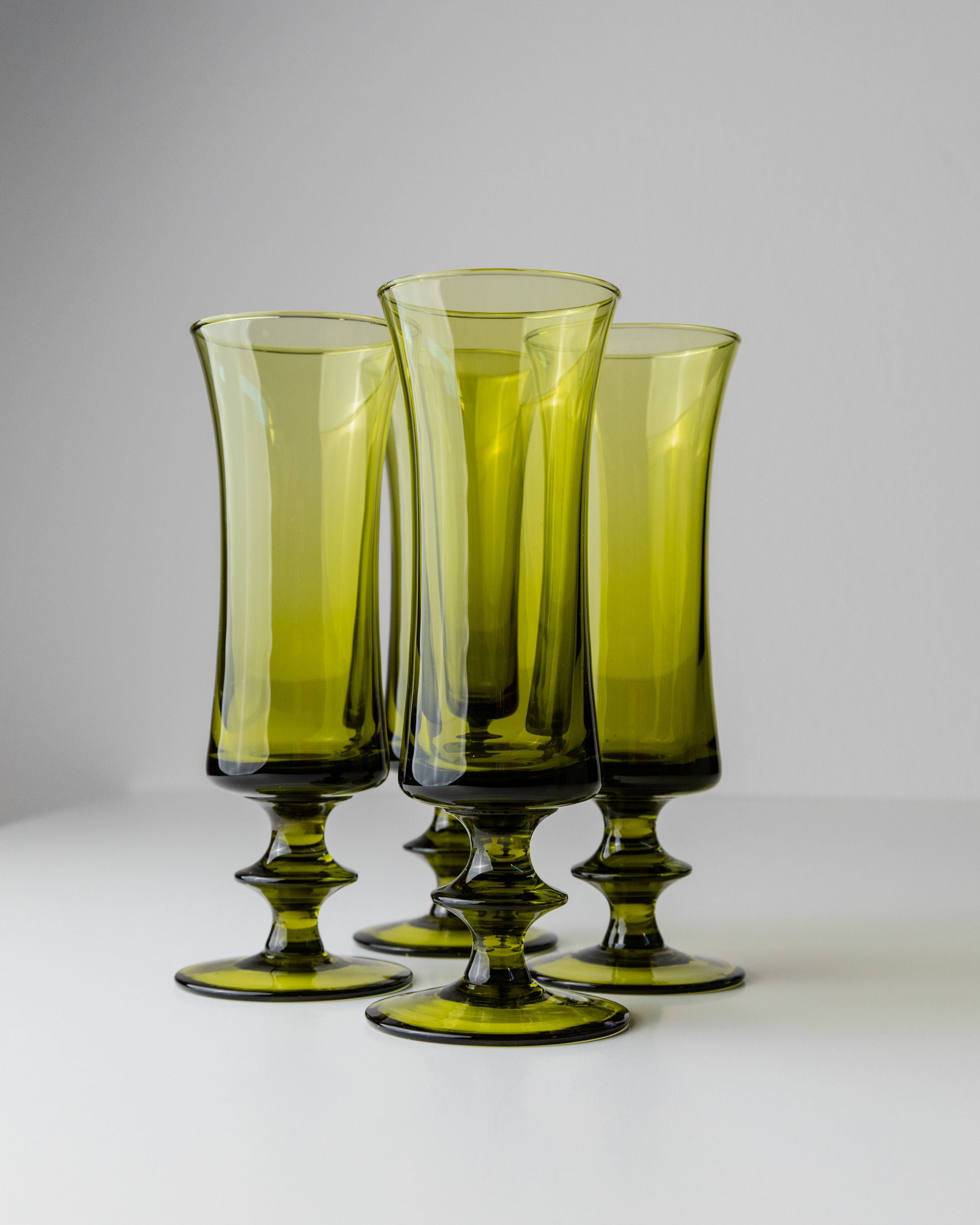 The 4 Murano Glasses in Green 70s by Bottega Jacobs, with flared rims and elegant stems, are closely arranged on a white surface against a gray backdrop, exuding mid-century design charm.