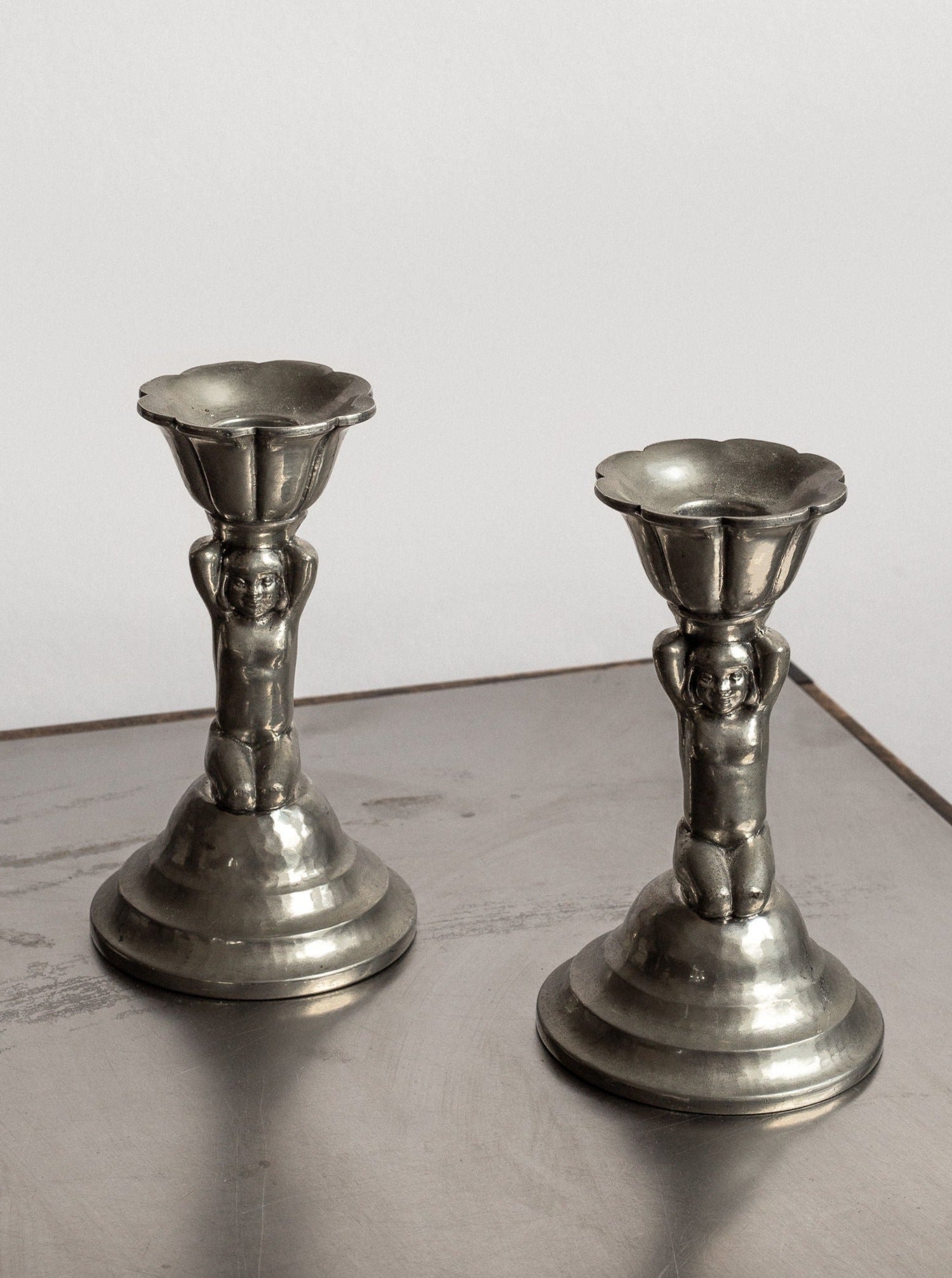 The 1930s Art Deco Metal Candlesticks from Spigel of Denmark each feature a small figure with outstretched arms, supporting a wide, fluted cup. The figures rest on tiered, circular bases against a smooth, plain backdrop. Their neoclassical design captures an air of timeless elegance.