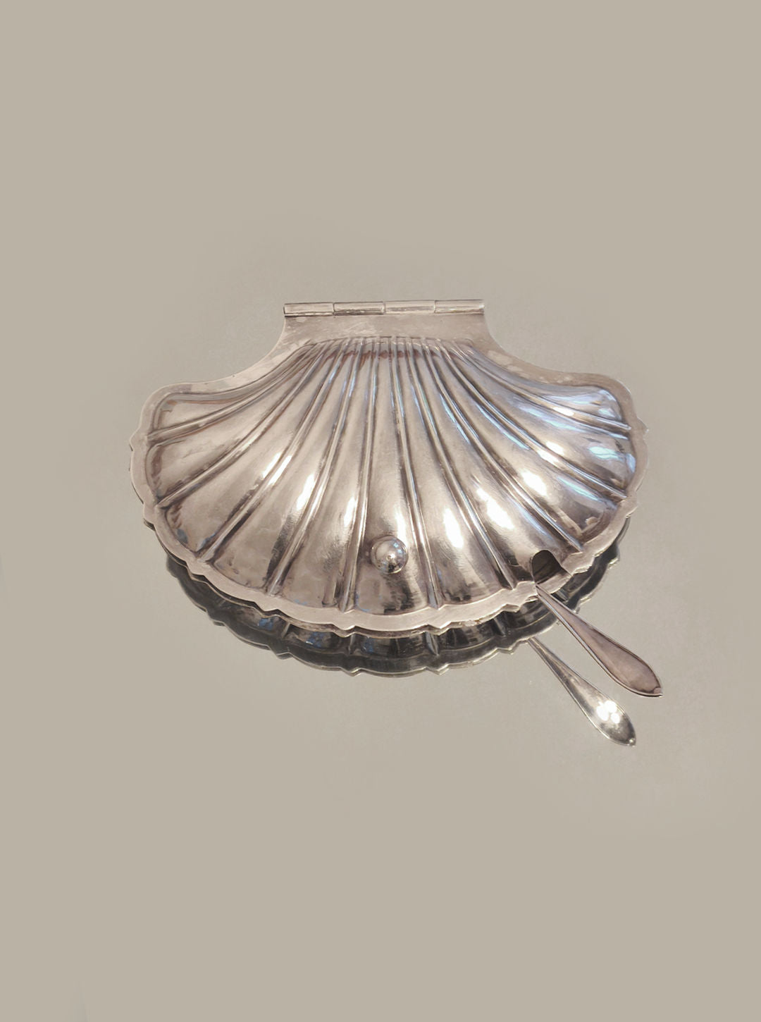 The Les Objoies Shell Butter Dish is a silver-plated piece with a shell-shaped design, featuring a hinged lid and small top handle. A matching spoon rests gracefully on the right, partly nestled inside. Its neutral gray background adds understated charm to refined dining settings.