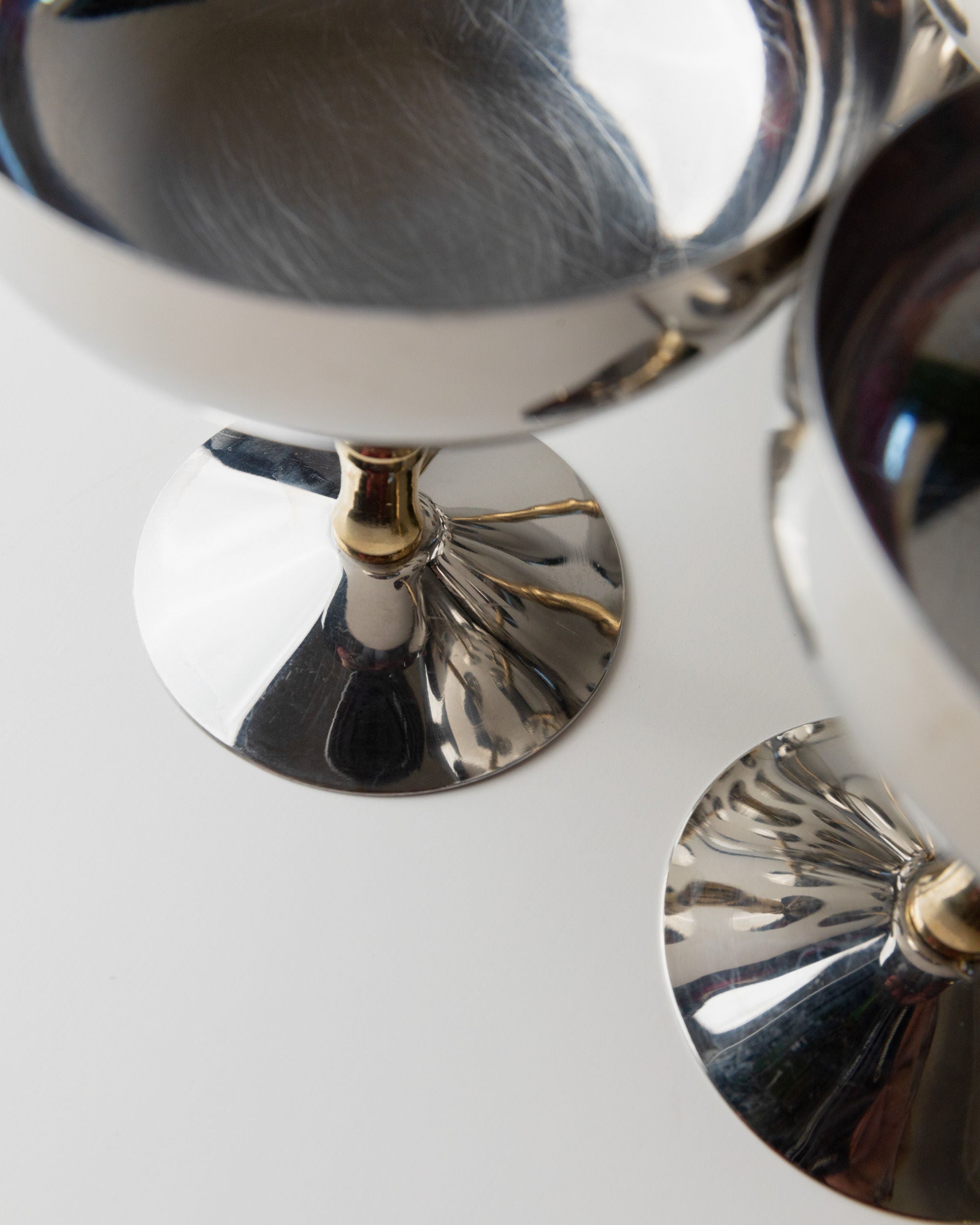 Close-up of two stainless steel champagne coupes from the 70s by Bottega Jacobs, featuring polished surfaces that reflect light and create intriguing reflections and distortions on a white surface.