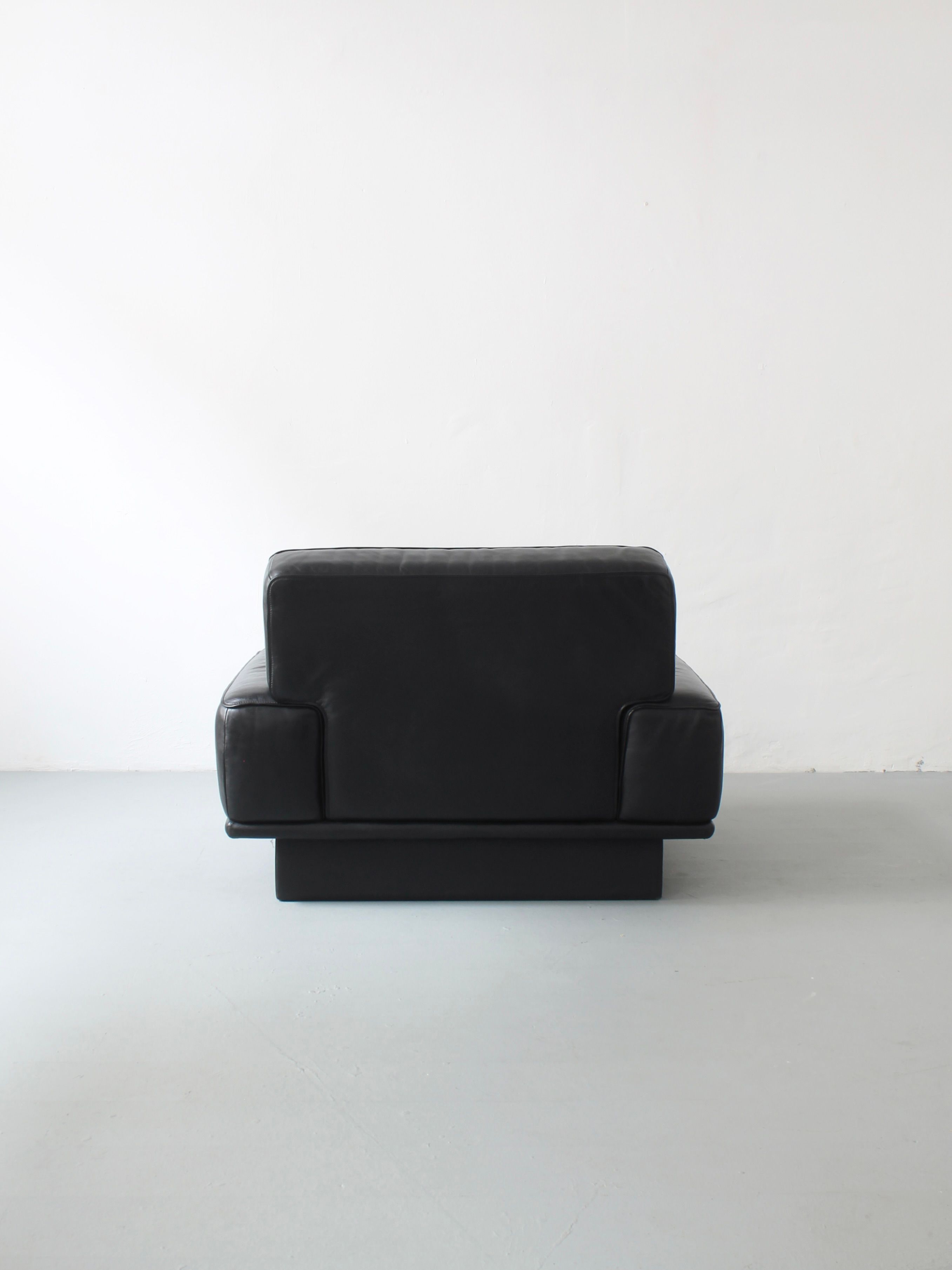 The black Lounge Chair by De Sede, from Afterhour Furniture, features a boxy design upholstered in high-quality leather. It sits against a plain white wall and light gray floor, exuding the sophisticated minimalist appeal of Swiss luxury furniture.