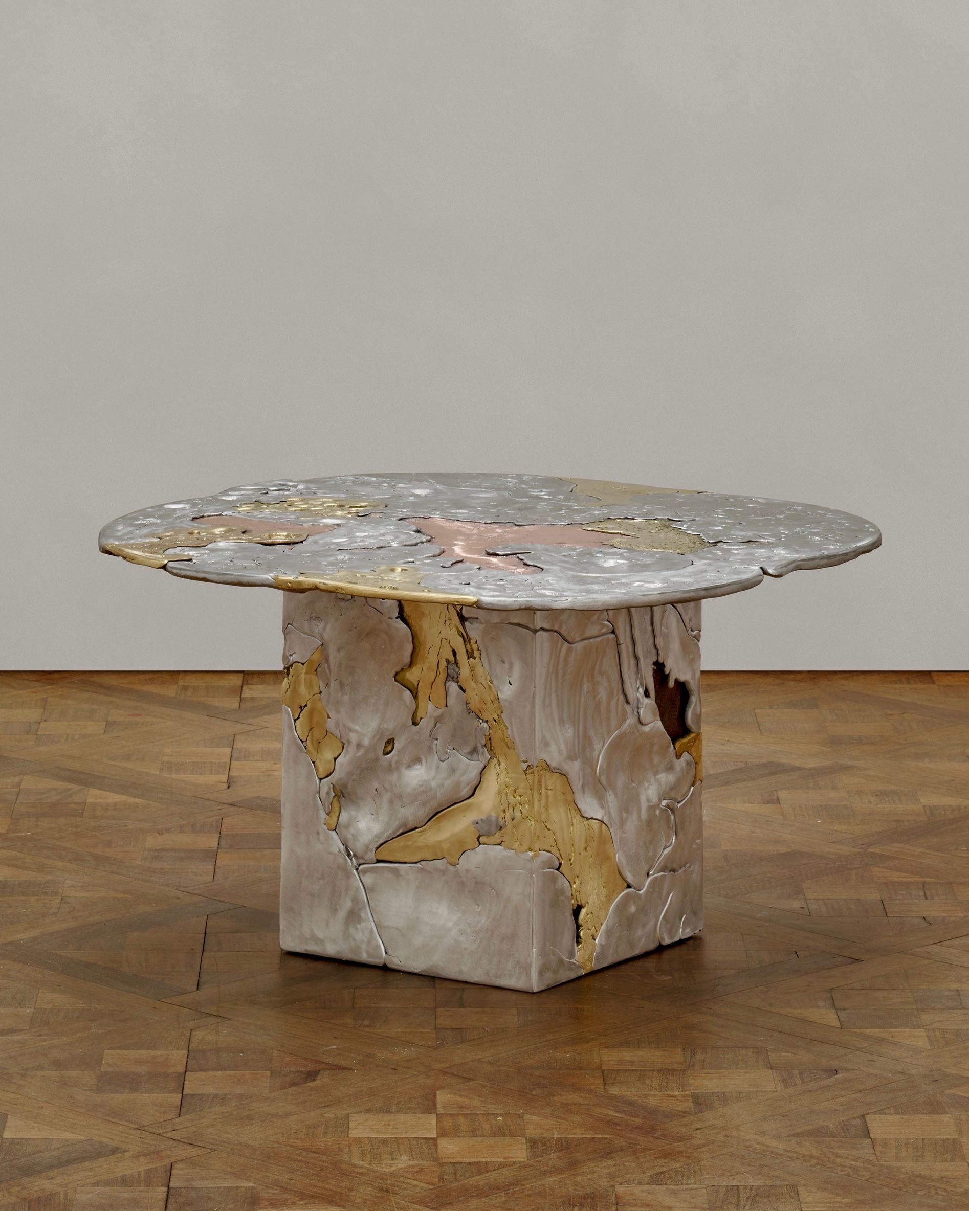 The 'Elemental Patchwork' Coffee Table by Sheyang Li features an irregular gray top with gold and pink marble accents, echoing molten metal, on a blocky crackled base. It stands on a wooden parquet floor against a plain wall.