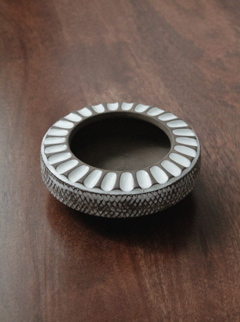 The Larholm Ceramic Ashtray Bowl by Vintage Stories is a round, shallow dish with a raised rim, white oval patterns, and a textured edge, shown here resting on a wooden surface.