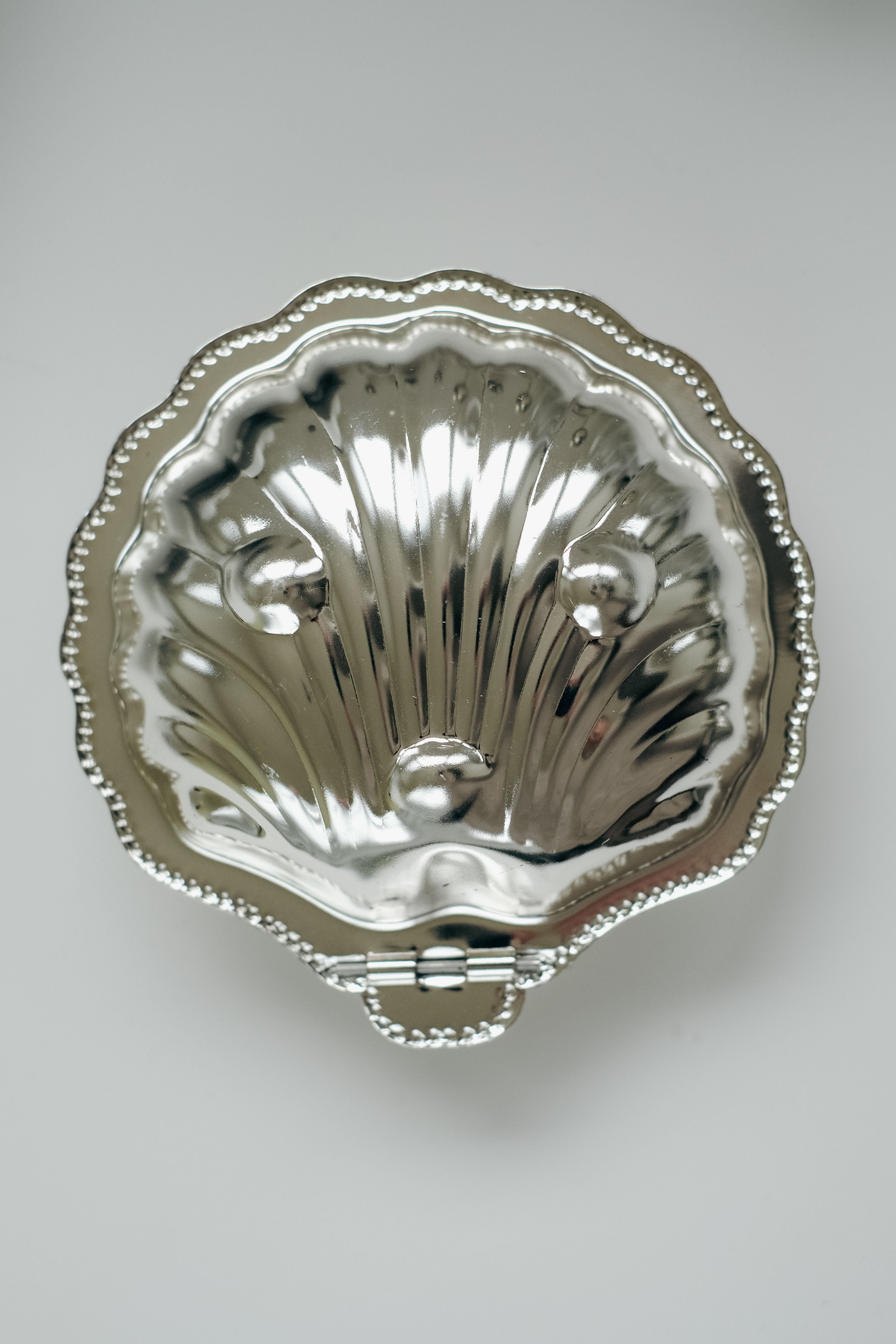 A silver trinket box from septembre studios, resembling the Shell Shaped Butter Dish, features a shiny clam shape with a detailed ridged design. Its hinged bottom opens to reveal an elegant shell pattern while resting on a white surface.