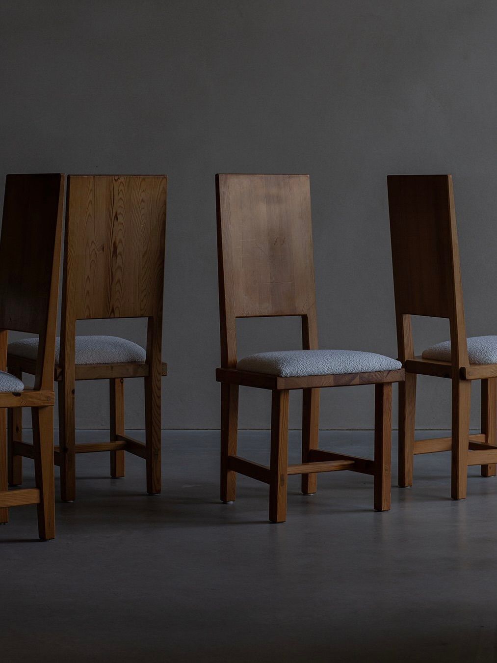 Four Model 553 chairs by Göran Malmvall for Bicci de' Medici, boasting tall rectangular backs and light gray cushioned seats, are arranged in a dimly lit room with a gray floor and background, exemplifying Swedish furniture design and positioned at varying angles.