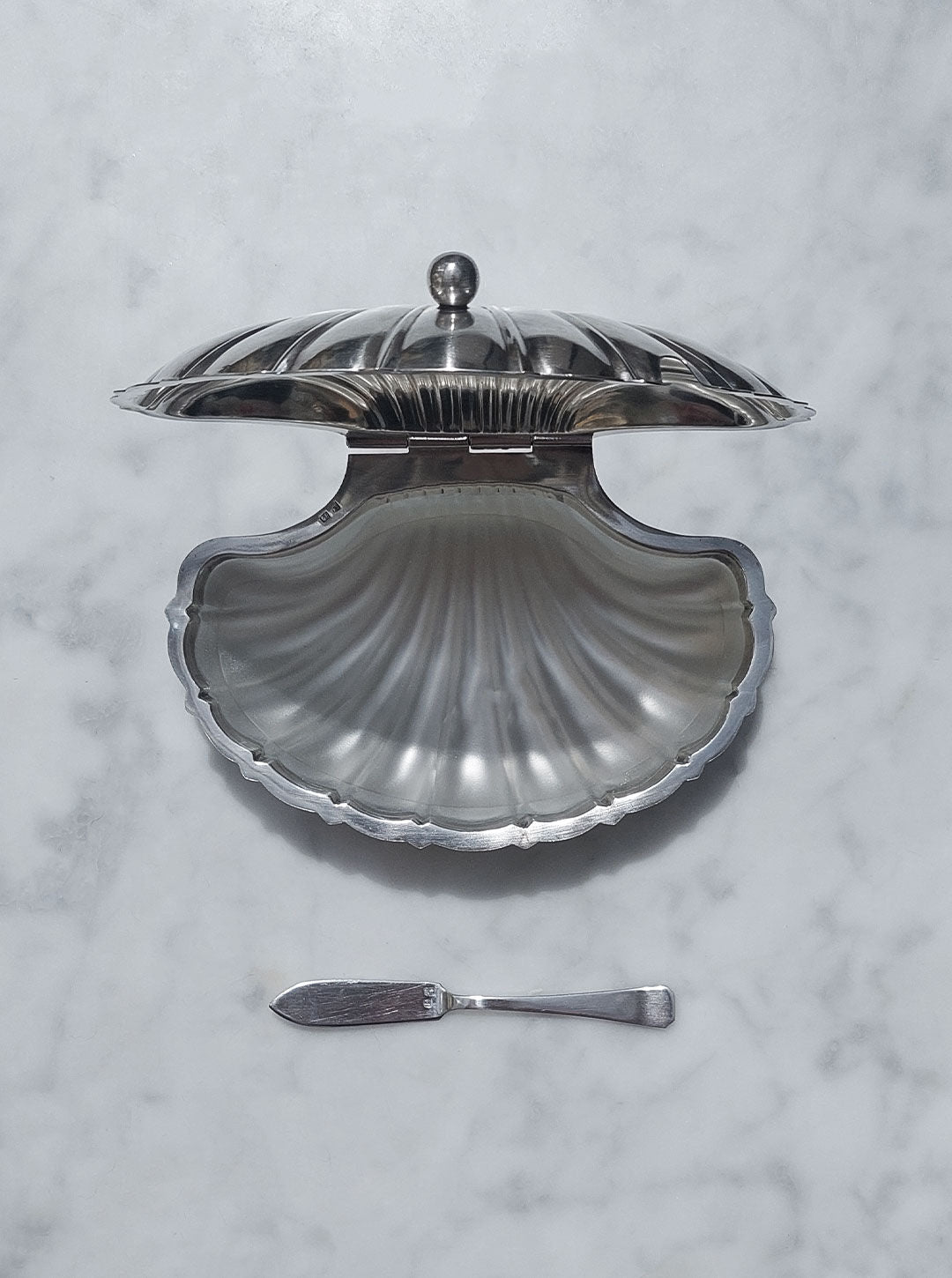 A Les Objoies Shell Silver-Plated Butter Dish with a hinged lid is open and empty, displayed on a white marbled surface, exuding vintage charm. Below the dish, there is a small, flat, silver-plated butter spreader with a simple design.