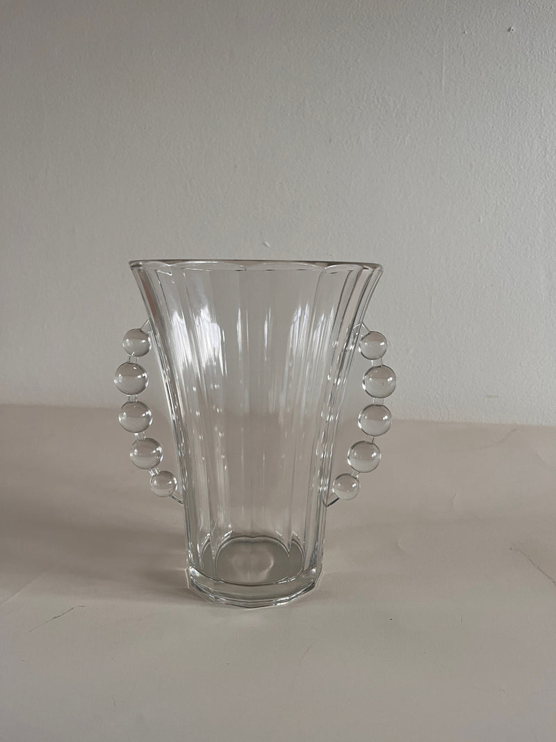 A Val Saint-Lambert vase by Maud Vaughan, featuring clear glass, fluted sides, and elegant beaded handles, sits on a plain surface against a neutral background.