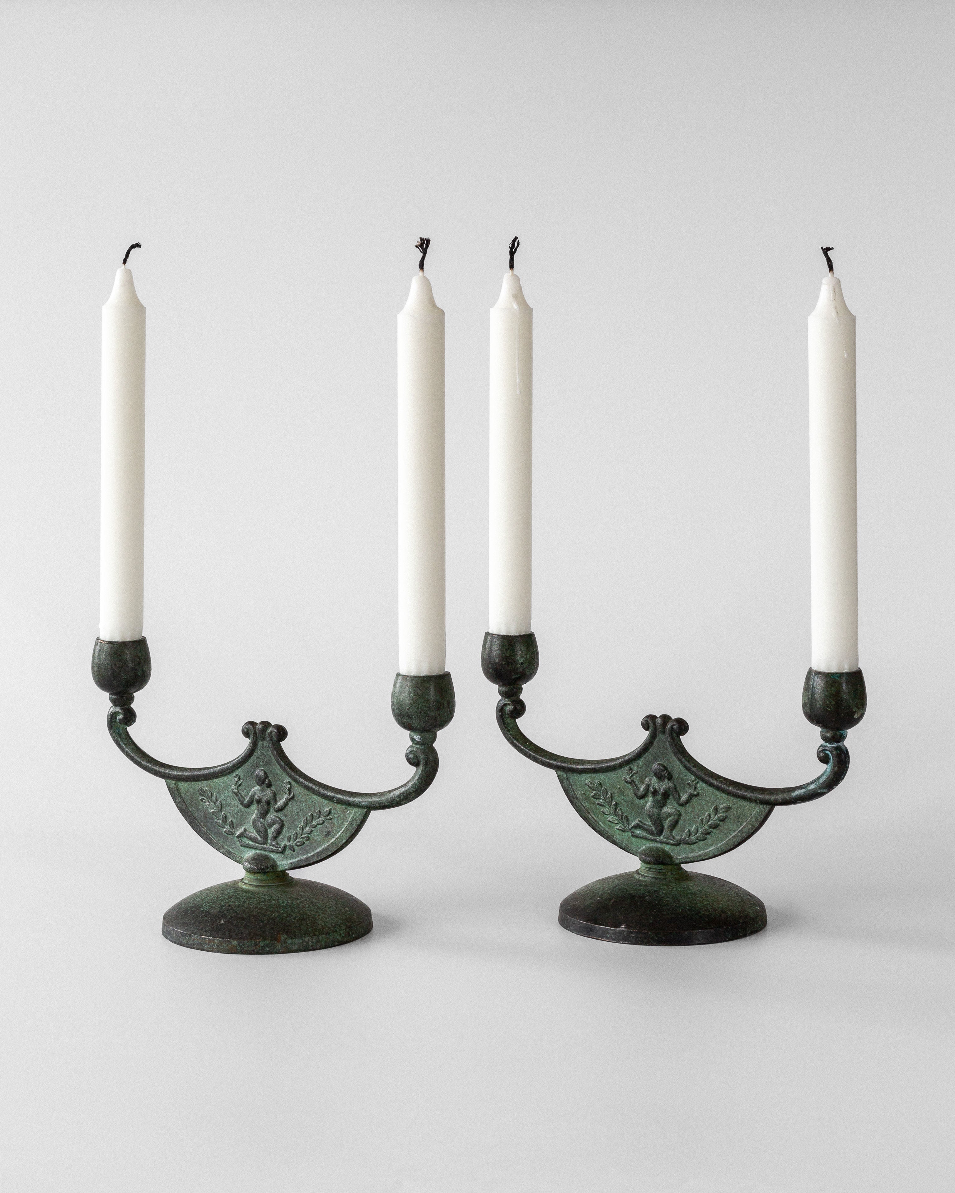 The Spigel Pair of Art Deco Bronze Candleholders, featuring exquisite Swedish metalwork and verdigris patina, each hold two unlit white candles against a plain white background.