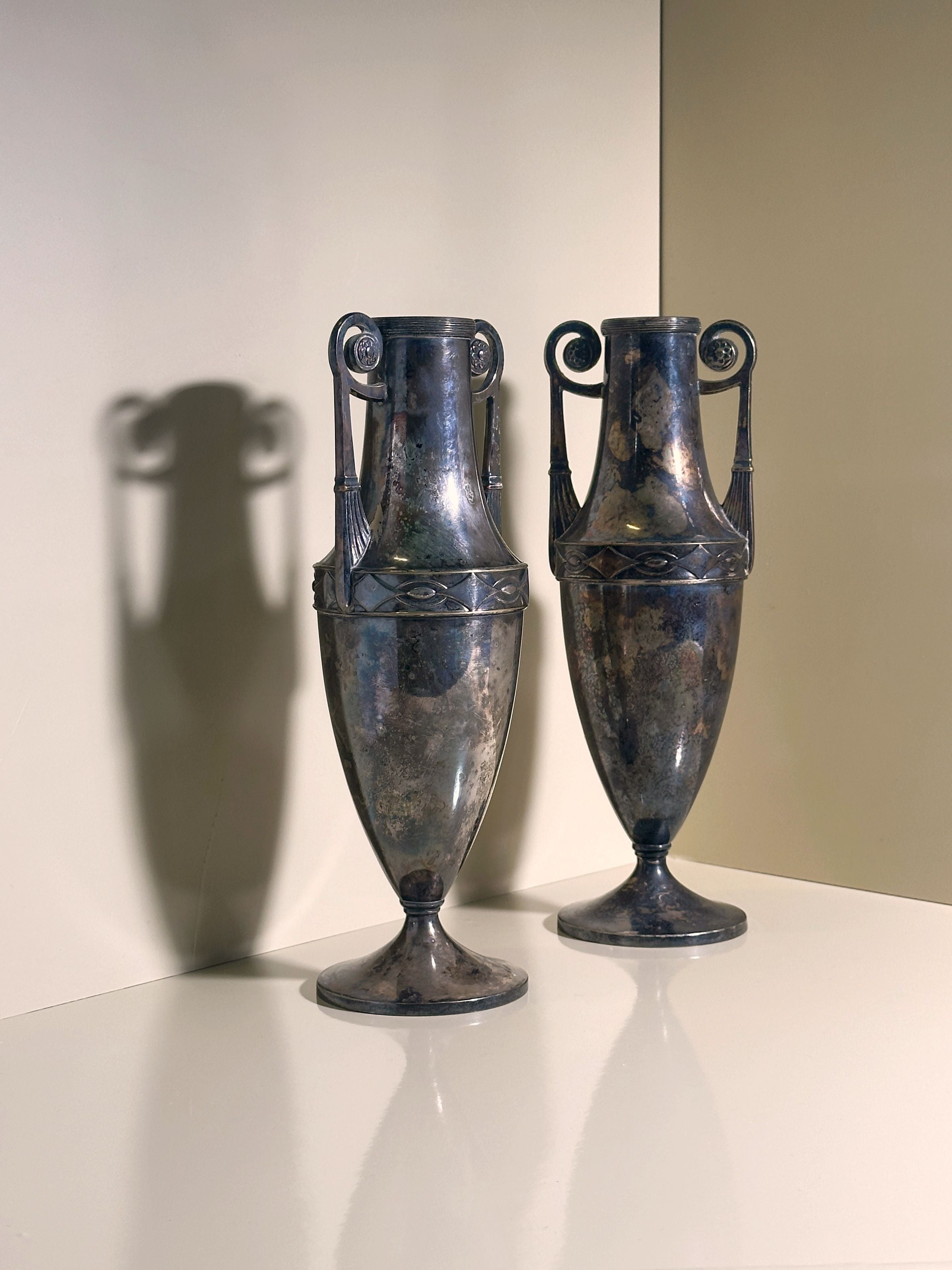 The Pair of Silver-plated Art Nouveau Amphora - 1890 by Dodo Vintage stands gleaming on a reflective surface, showcasing ornate handles. They cast soft shadows against a neutral backdrop, with subtle reflections on the surface below.