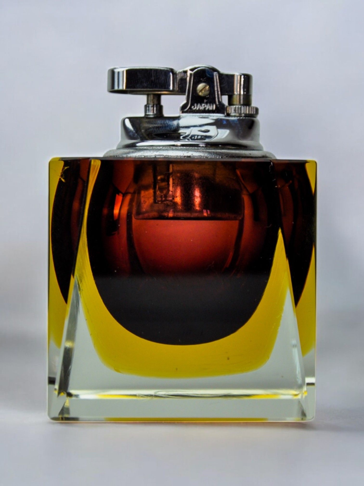 The Sommerso Lighter by Seguso from Malamar Studio features a square, transparent Murano glass bottle partly filled with amber liquid and topped with a shiny silver mechanism. This elegant piece from the 70s echoes Flavio Poli's vintage designs against a plain background.