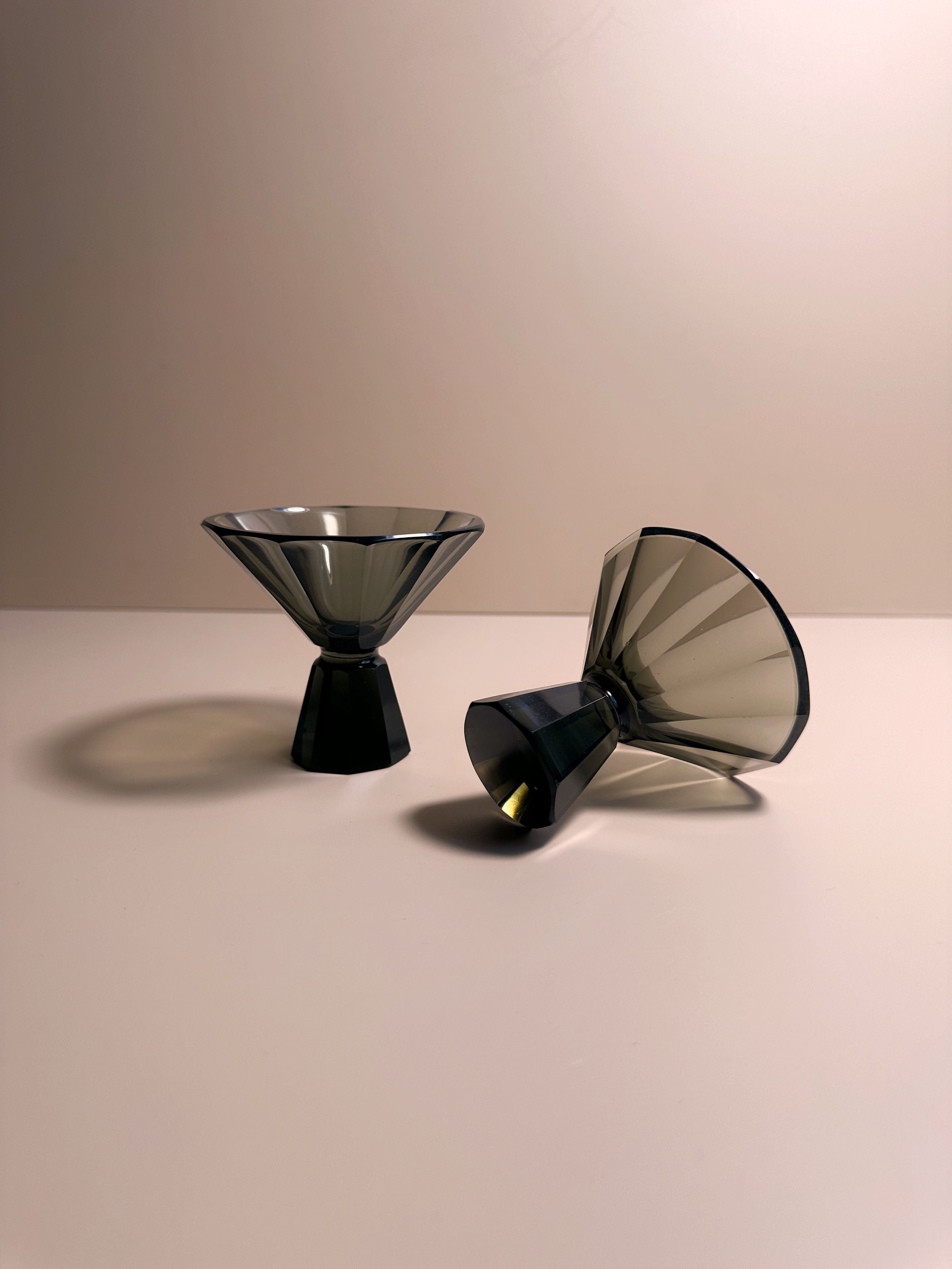 Two handmade 1930s Art Deco Facetted Liqueur Glasses from Dodo Vintage, featuring inverted pyramid shapes with dark bases, are arranged on a smooth, light-colored surface. One smoked glass is upright while the other rests on its side. The background is plain and softly lit, creating delicate shadows behind the objects.