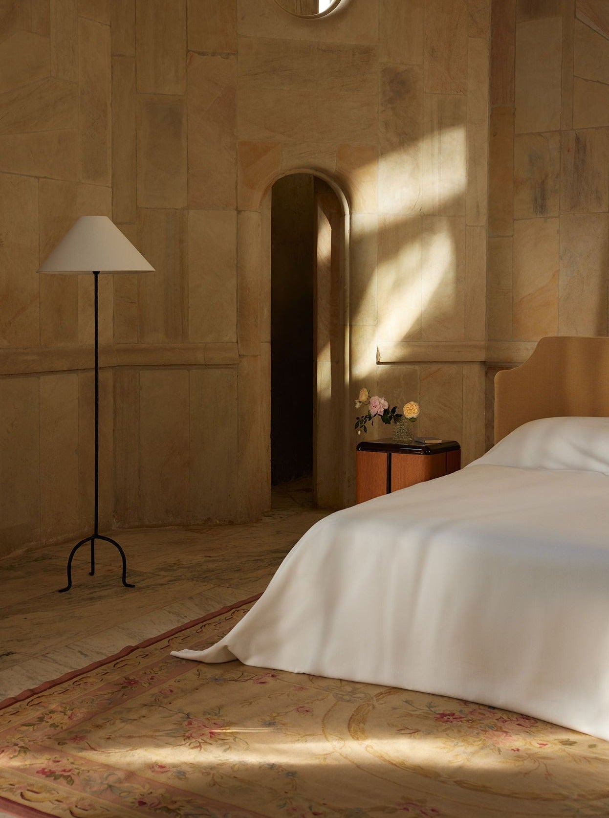 A serene bedroom with sunlight streaming through a tall window, casting a warm glow on a beige wall. The room features an elegantly draped bed, a standing lamp, and a Lemon Pair of Various Positions Nightstands.