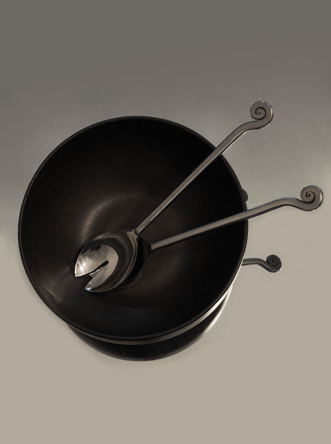 The Les Objoies Spiral Salad Servers, paired with a black bowl, feature sculptural handles with decorative spirals, adding charm against a neutral gray backdrop.