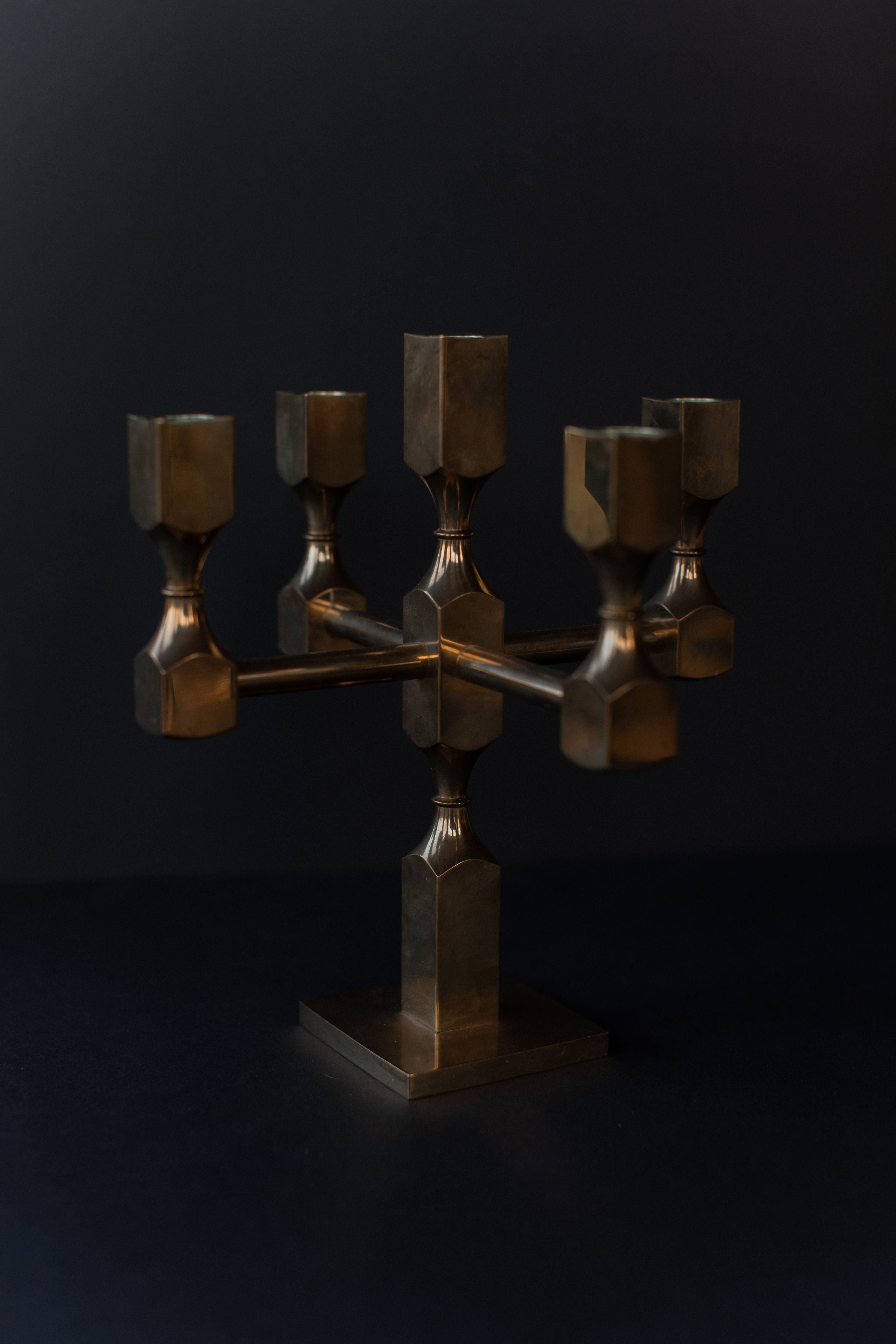 The Brutalist Brass Candleholder by Out For Lunch features a geometric design with square holders on a sturdy base, embodying an industrial aesthetic in bronze against a dark background.