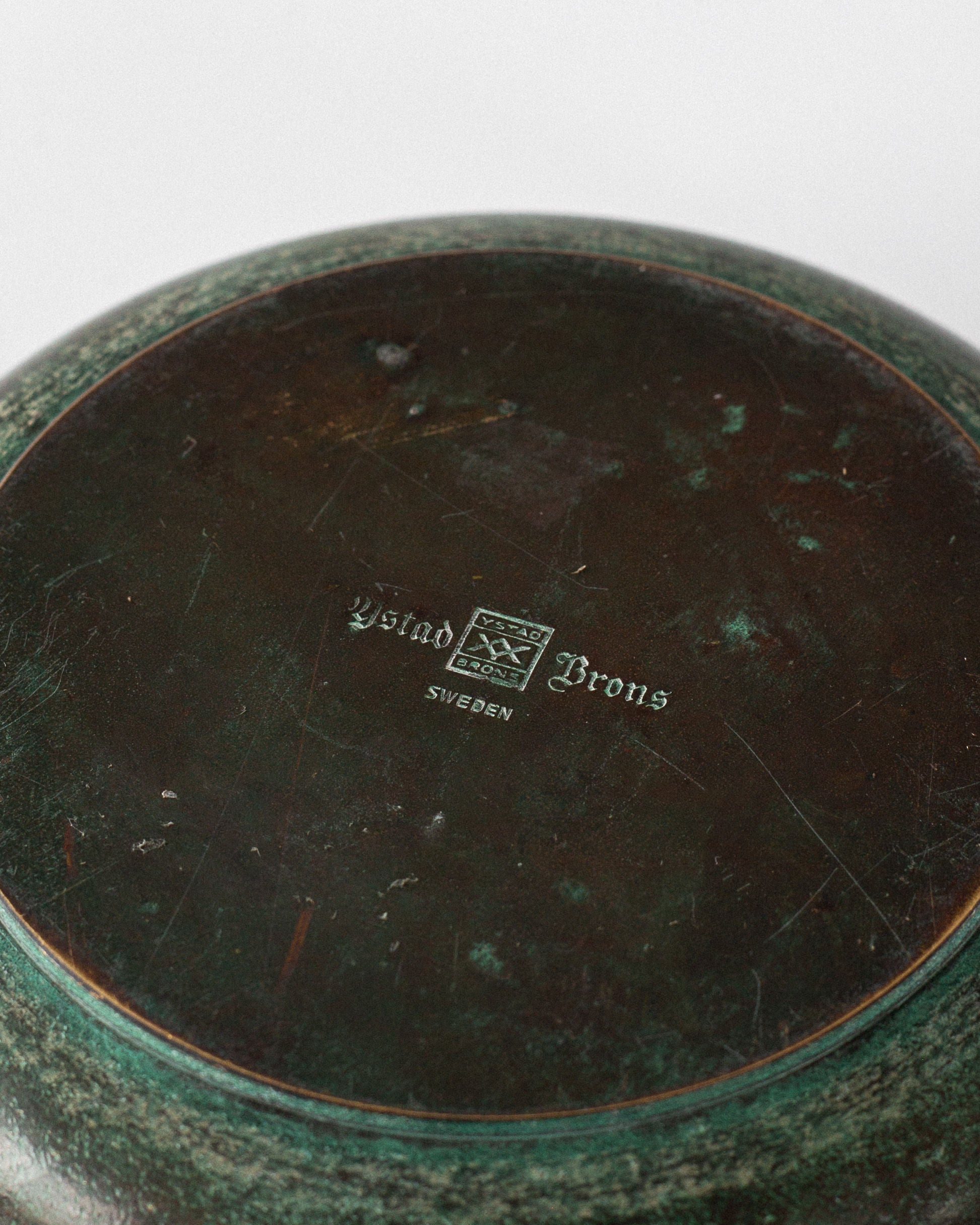 The image displays the underside of a weathered, round Art Deco bronze bowl by Spigel from Sweden, 1930s. It features a greenish patina with scratches and a central logo engraved with "Ystad Brons Sweden," echoing classic designs like those with Greek key handles.