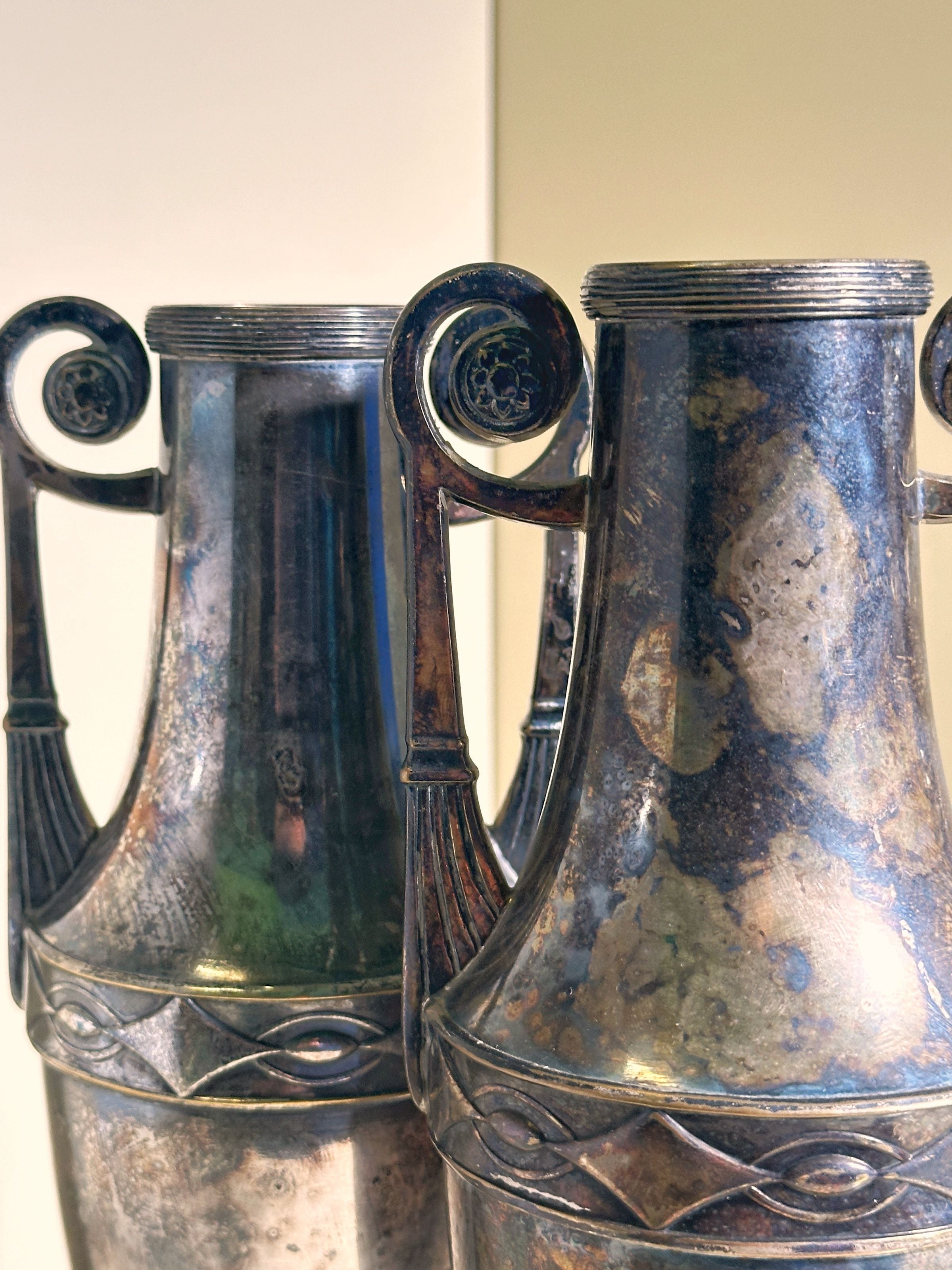 Positioned closely together, the Dodo Vintage Pair of Silver-plated Art Nouveau Amphora from 1890 feature intricate, curved handles and a weathered, mottled patina that enhances their antique charm.