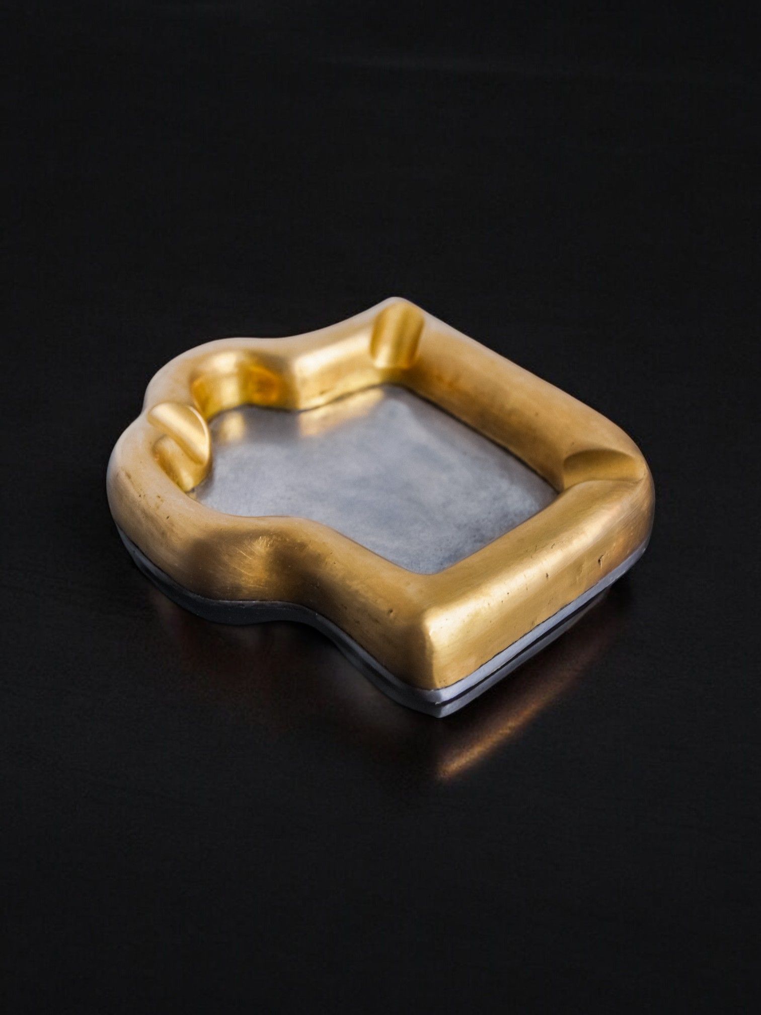 A rectangular, bronze and aluminum "Puzzle" ashtray by David Marshall from 1970, featuring curved edges, is elegantly displayed on a black surface. Offered by Malamar Studio.