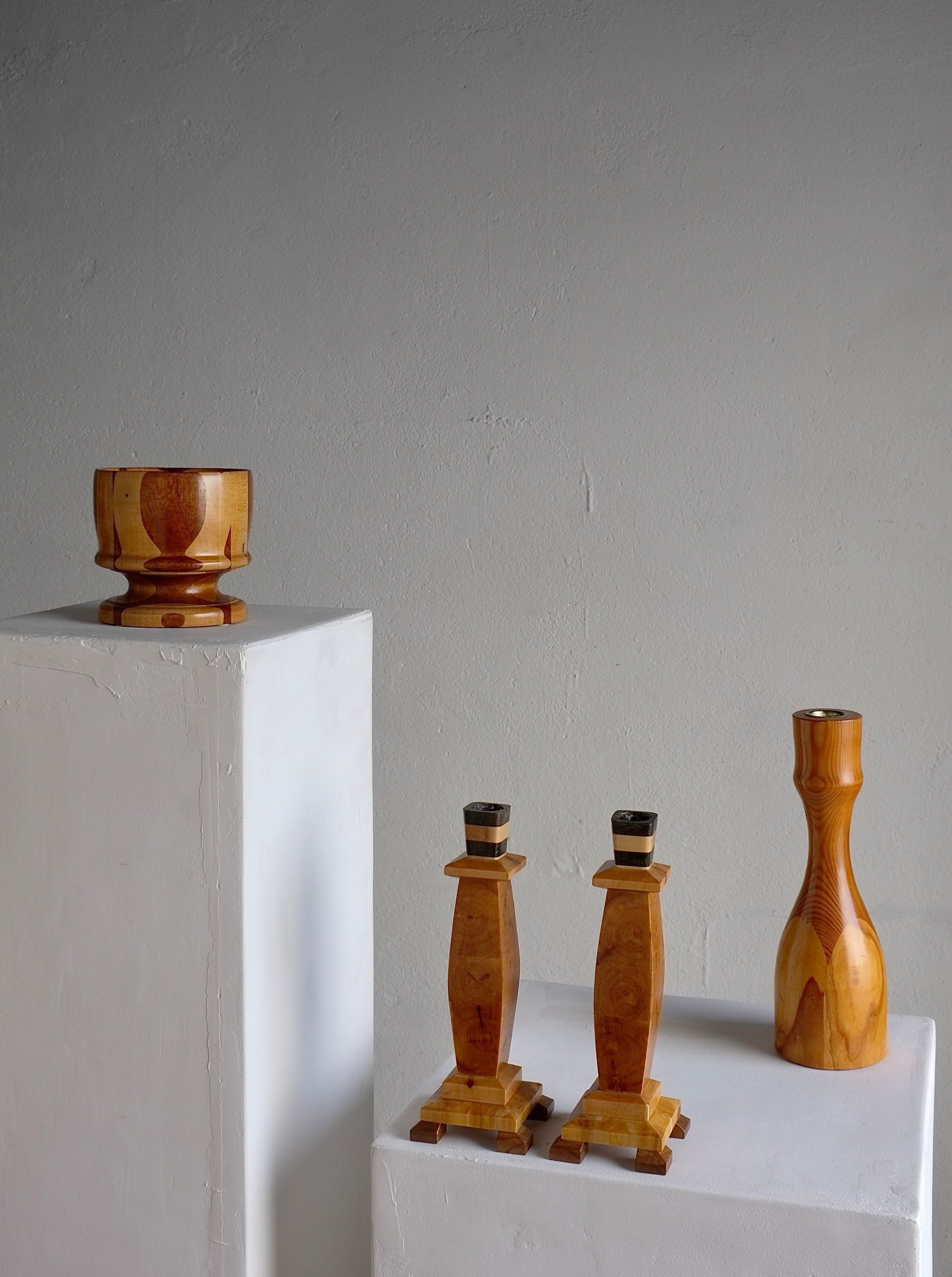 A minimalist composition showcasing vintage carved wooden decor on white pedestals. On the left, a small wooden bowl from the 1970s, part of Veter Vintage's Carved Wooden Candle Holders and Bowl Set, sits atop a tall pedestal. The right pedestal features two wooden candlestick holders along with a tall, slender wooden vase. The background is a simple, neutral wall.