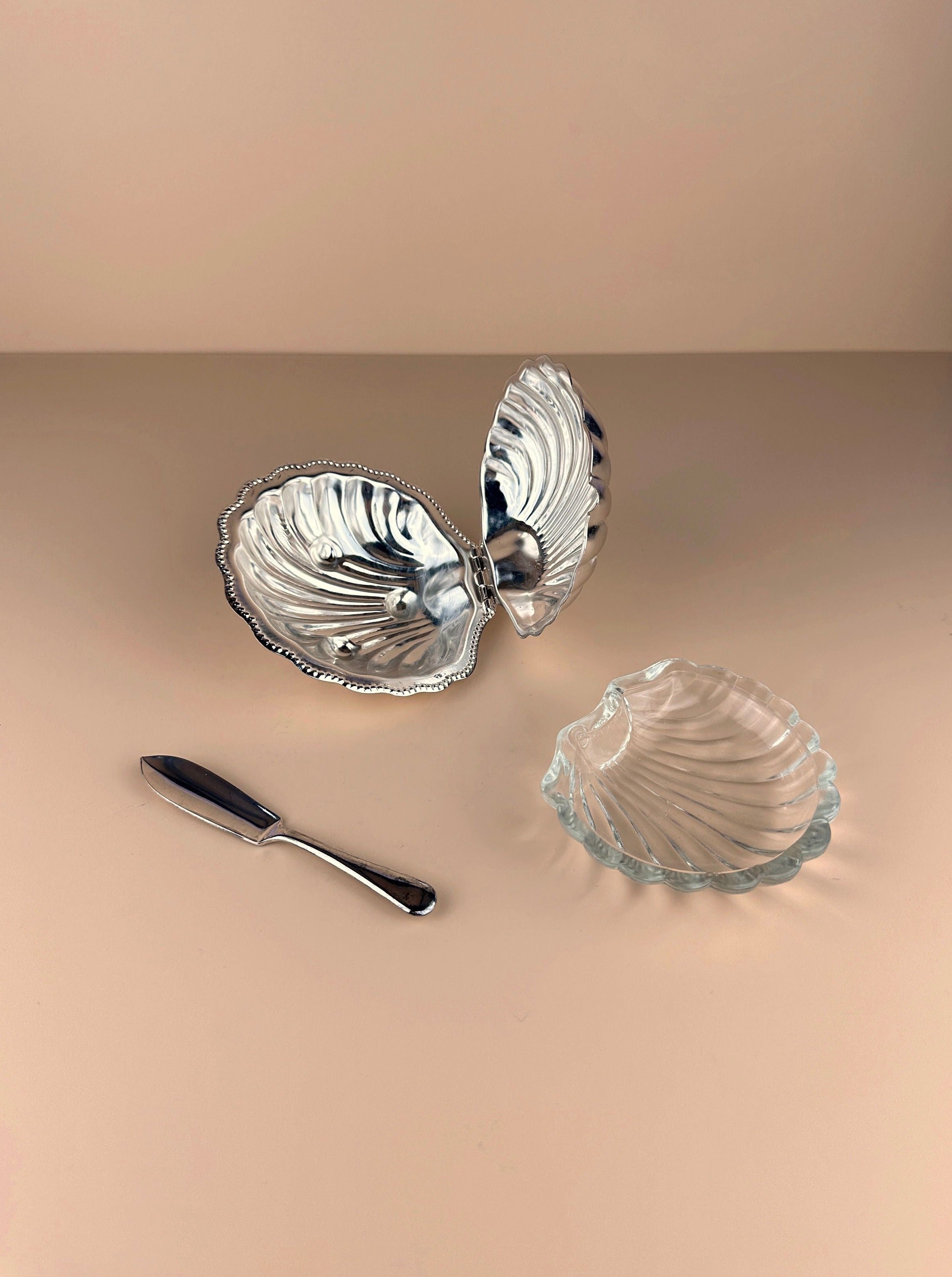 An open, Dodo Vintage Shell Shaped Silver Plated Butter Dish with intricate designs sits on a beige surface. Next to it is a matching glass dish and a small silver butter knife. The setup appears elegant and minimalist against the neutral background.