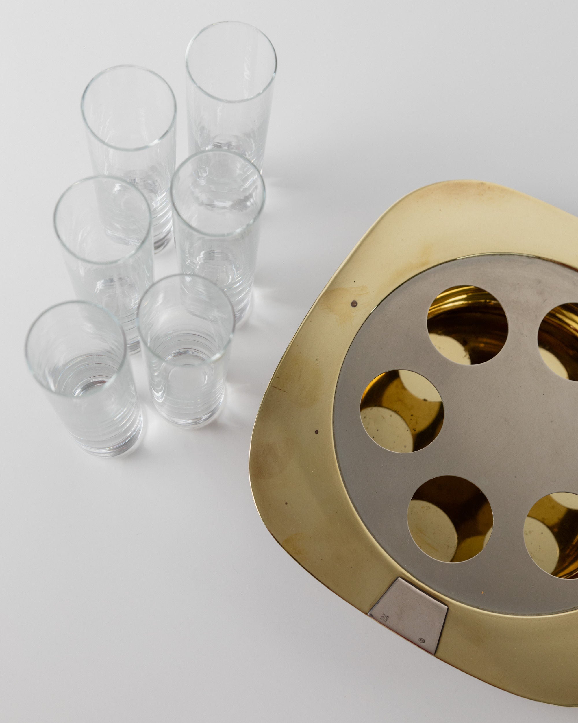 A set of seven vintage shot glasses from Bottega Jacobs' Set Shot Glasses and Tray - Mid Century Design 70s rests on a white surface beside a gold-colored tray with circular cutouts, boasting a shiny metallic finish that adds a decorative mid-century accent.