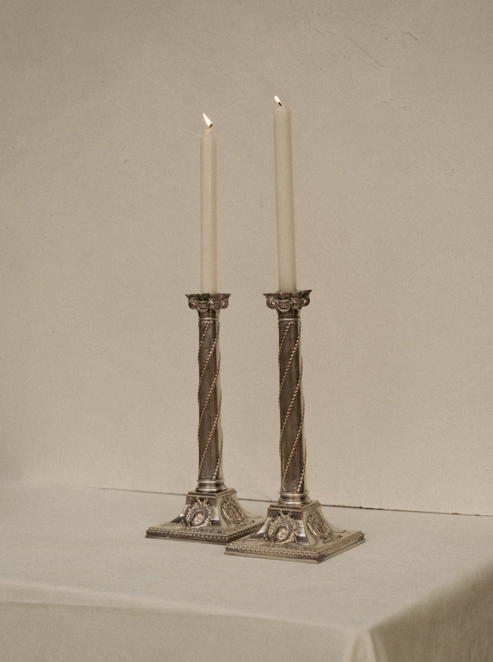 Two lit tall white candles stand in ornate Casita Amada Pair of Column Sheffield silver plated candleholders. The antique holders, with intricate designs and twisted, rope-like embellishments, rest on a white table against a neutral beige background. The detailed square bases add a touch of timeless elegance.