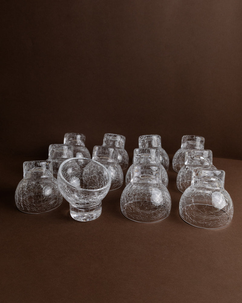 A set of 12 wine and cocktail glasses from the Bottega Jacobs brand, originating in Italy during the 1970s, are arranged on a brown surface. These transparent glasses capture and reflect light, producing delicate highlights against the dark background, evoking the elegance of mid-century design.