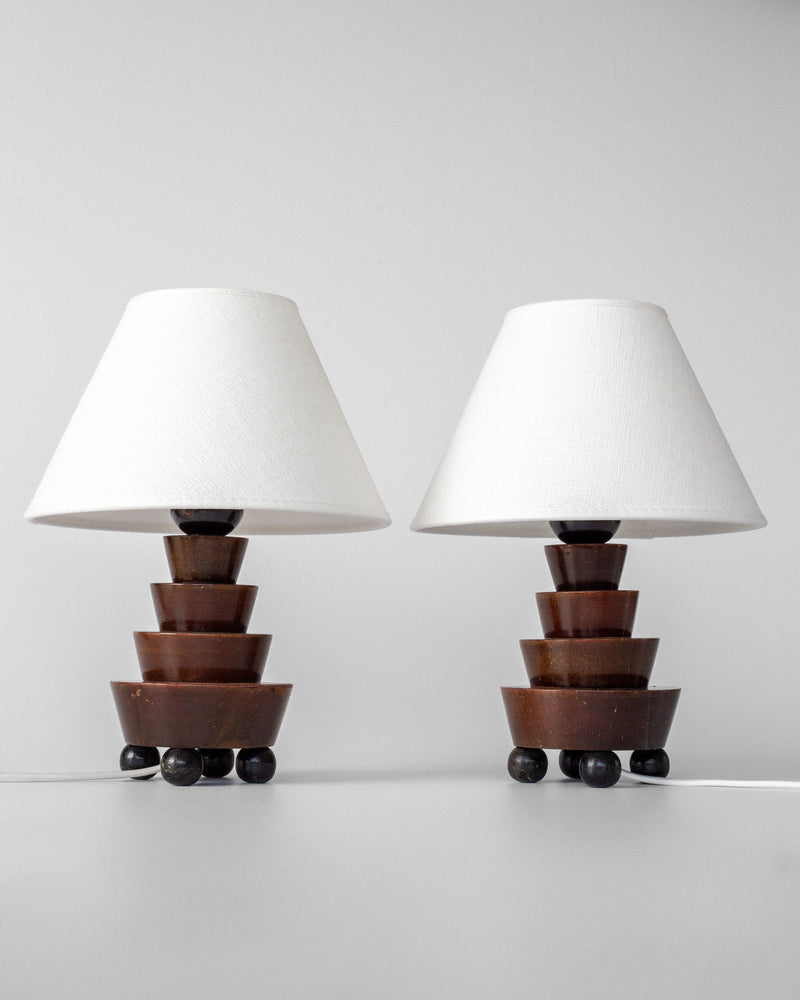 Two Spigel Art Deco Wooden Table Lamps with white cone-shaped shades and tiered geometric brown bases, each on four black spherical feet, are displayed against a plain light gray background.