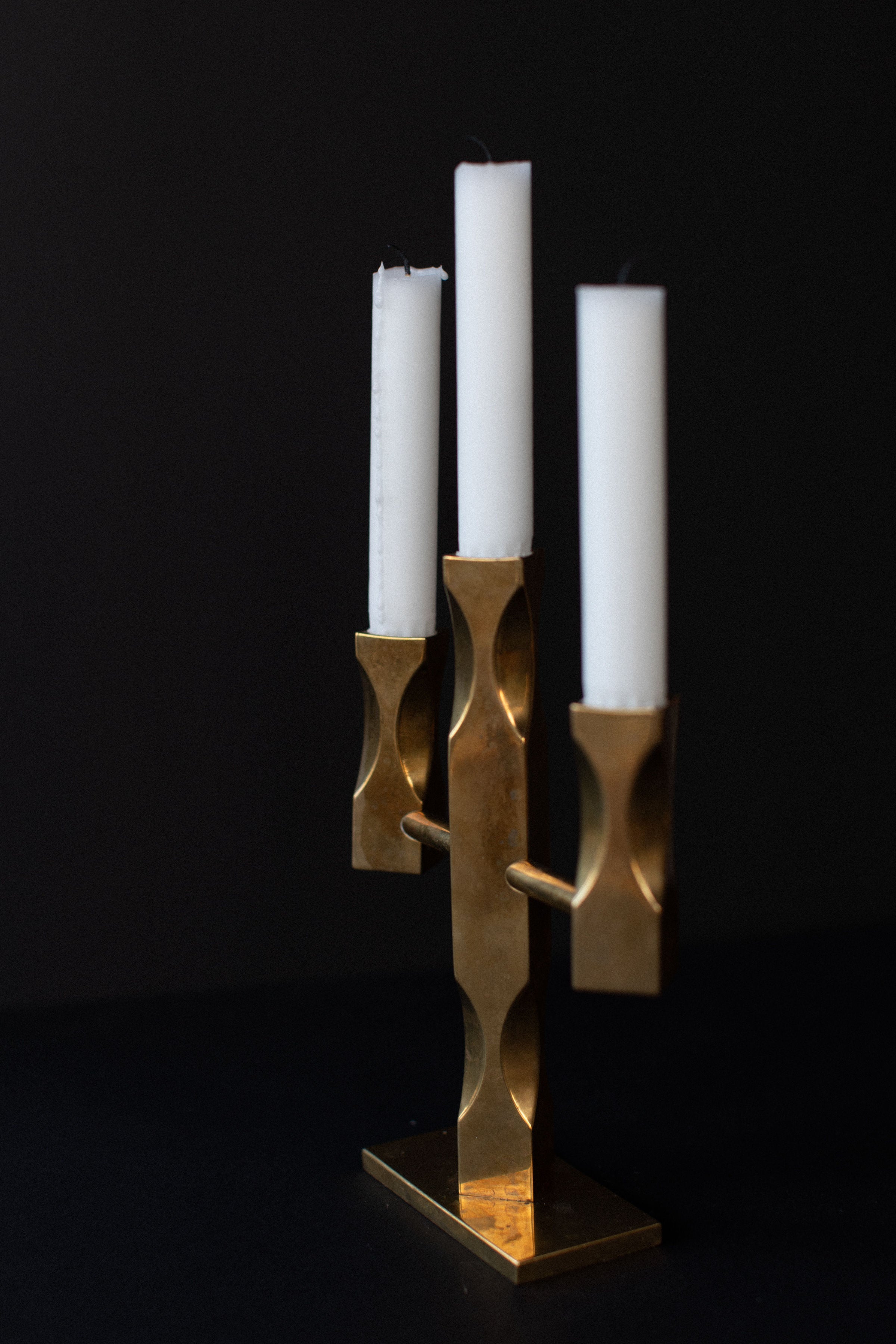 The Out For Lunch Brass Candleholder is a tall, elegant Brutalist piece in solid brass holding three white candles against a dark background. It features a modern geometric design with smooth, rounded curves that create a captivating sculptural form.