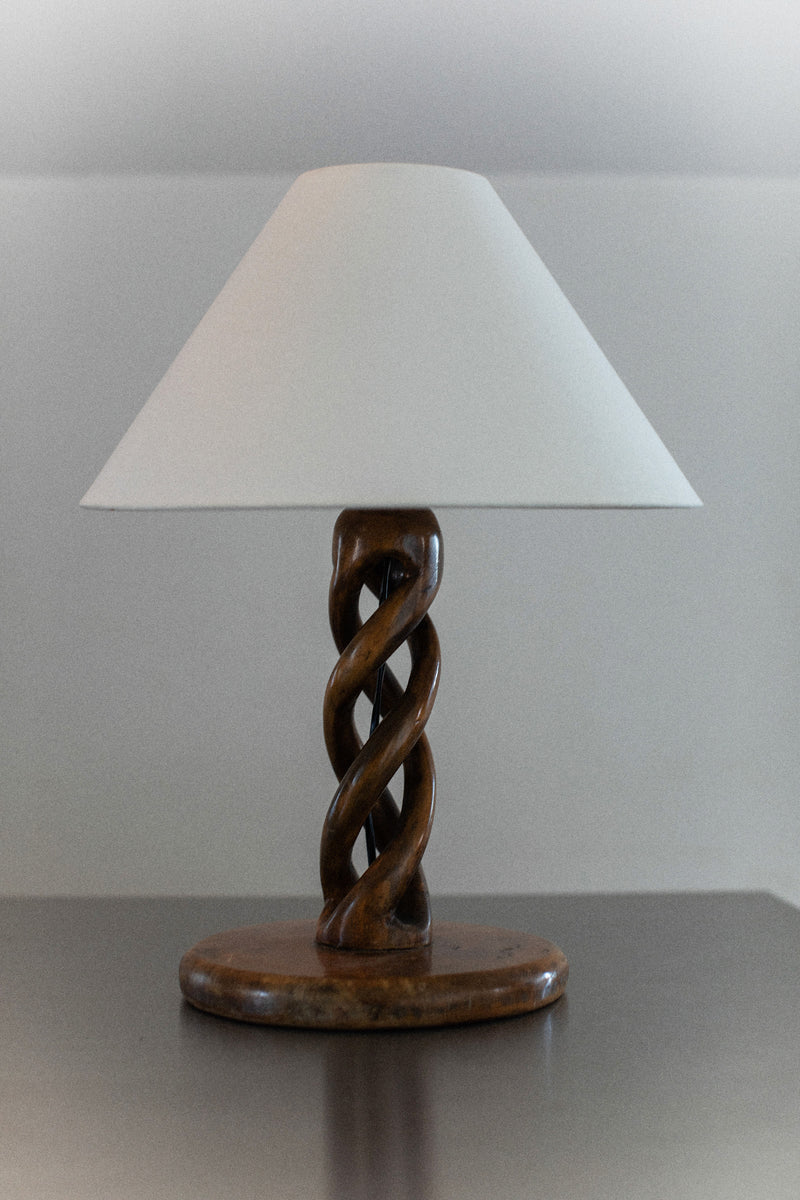 The Early-Century Lamp by Out For Lunch, featuring a white conical shade, is displayed on a dark surface. Its base mimics a double helix shape with intricately carved twisted wooden details, bringing an artistic flair to the minimalist setting.