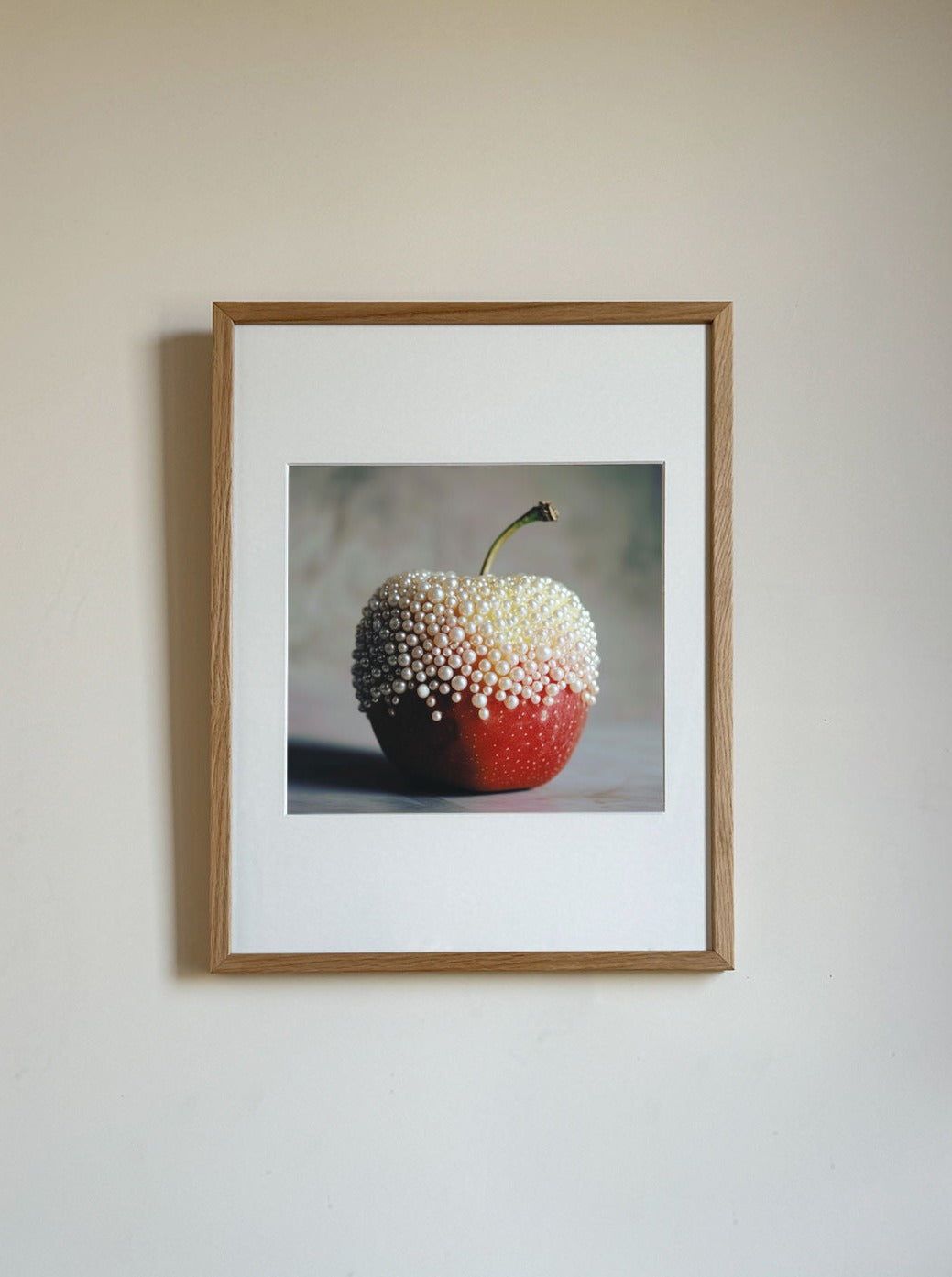 A fine art print of Apple and Pearls by Adeline Mai, covered in tiny white beads, hangs on a plain off-white wall. The simple frame has a light wooden finish, and the limited edition image is centered within a white matting.