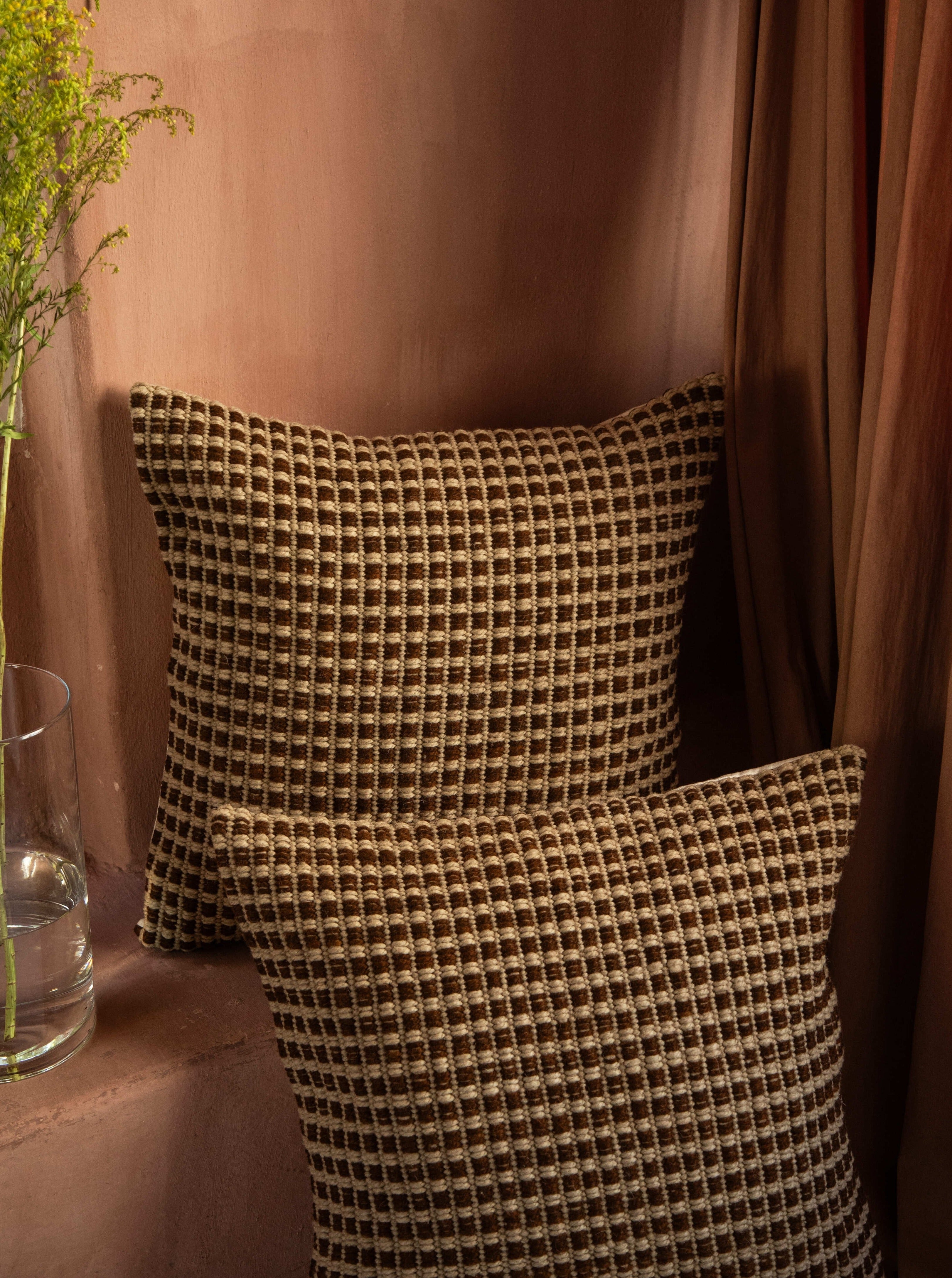 Two Cima Pillows from Identidad Argentina made from hand-spun sheep wool in a warm, cozy corner with ambient lighting, draped curtains, and a hint of greenery from a small plant.