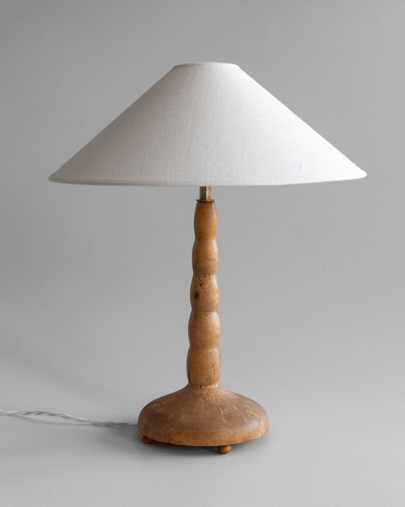 The Spigel Turned Birch Table Lamp from Sweden features a textured wooden rounded base, smooth birch stem, brass fixture, and large white conical lampshade, resting on a neutral background with a visible power cord.