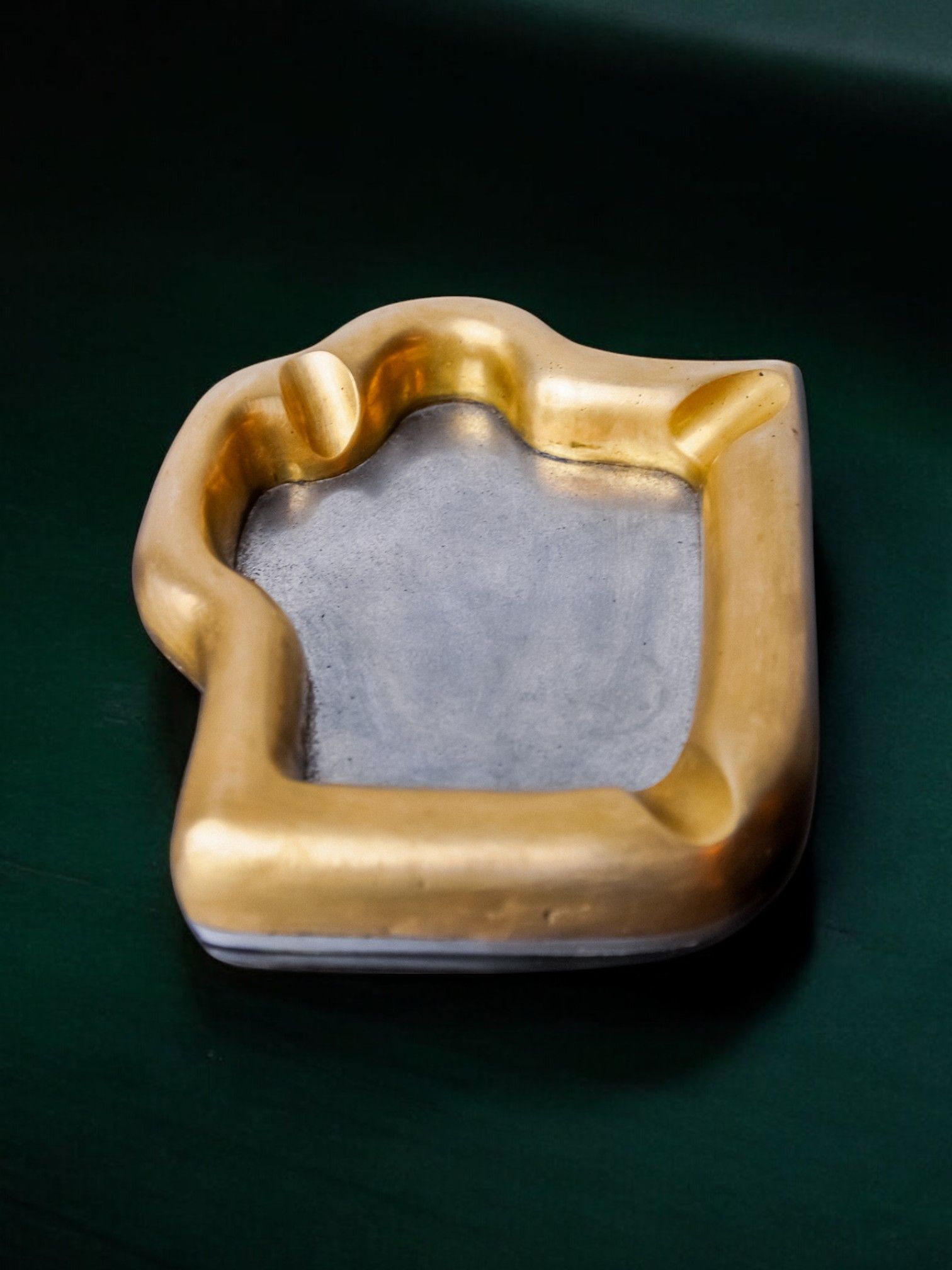 A Brutalist "Puzzle" Ashtray by David Marshall, created in Spain in 1970 and available at Malamar Studio, is a gold-trimmed ceramic piece with an irregular shape and smooth gray center, resting on a dark green surface.