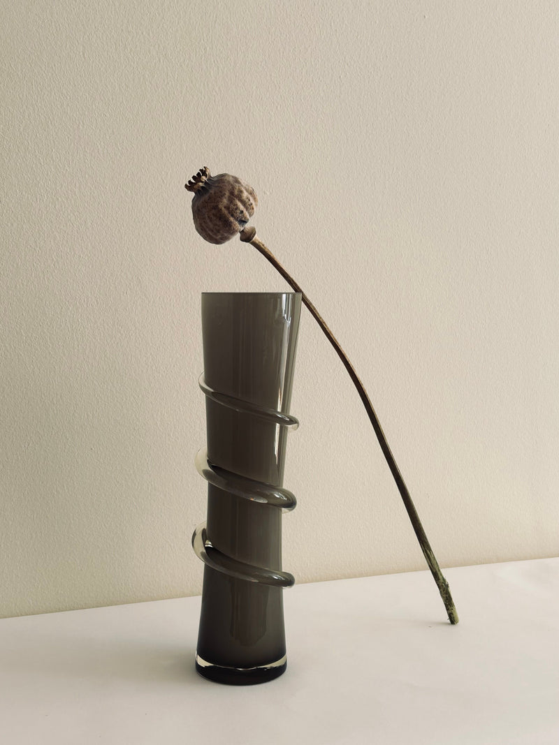 The Glass Vase with Spiral Detail by Flock Studio showcases a single dried flower stem accompanied by a seed pod. This minimalist composition, featuring the tall smoky glass vase with its unique spiral wrap detail, is elegantly set against a plain, light-colored background.