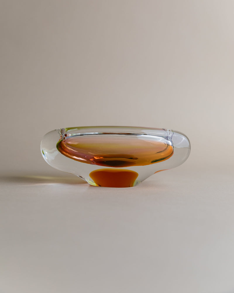 The Glass ashtray by Josef Rozinek for Novy Bor, from Bottega Jacobs, has a unique rounded design with an amber interior. Its transparent material reflects light, creating a gradient effect on the neutral surface it sits on.
