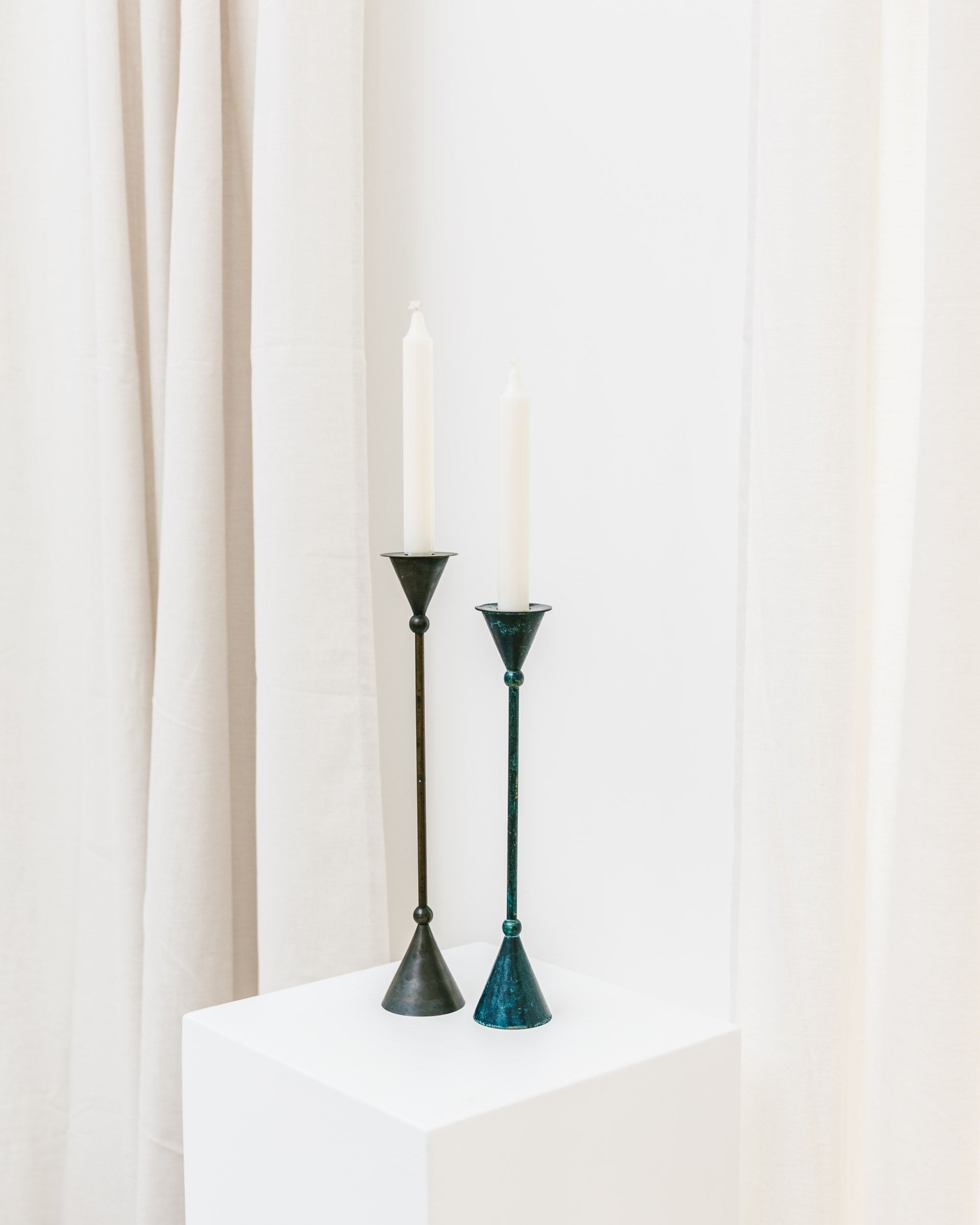 The Set of 2 Candleholders – Postmodern 1980s from Bottega Jacobs showcases tall, slender designs reminiscent of vintage postmodern style. Featuring white candles on a white pedestal, the conical form enhances their elegance against light beige drapes, creating a minimalist and refined setting.