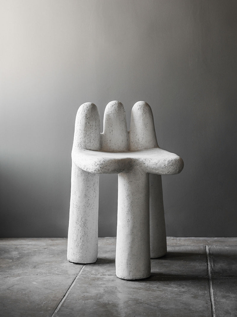 The Washed Concrete Chair by Studio OSKLO features a white, textured finish with three rounded legs and an abstract, organic form. Handmade from molded concrete, this unique piece stands out against a minimalist gray background.