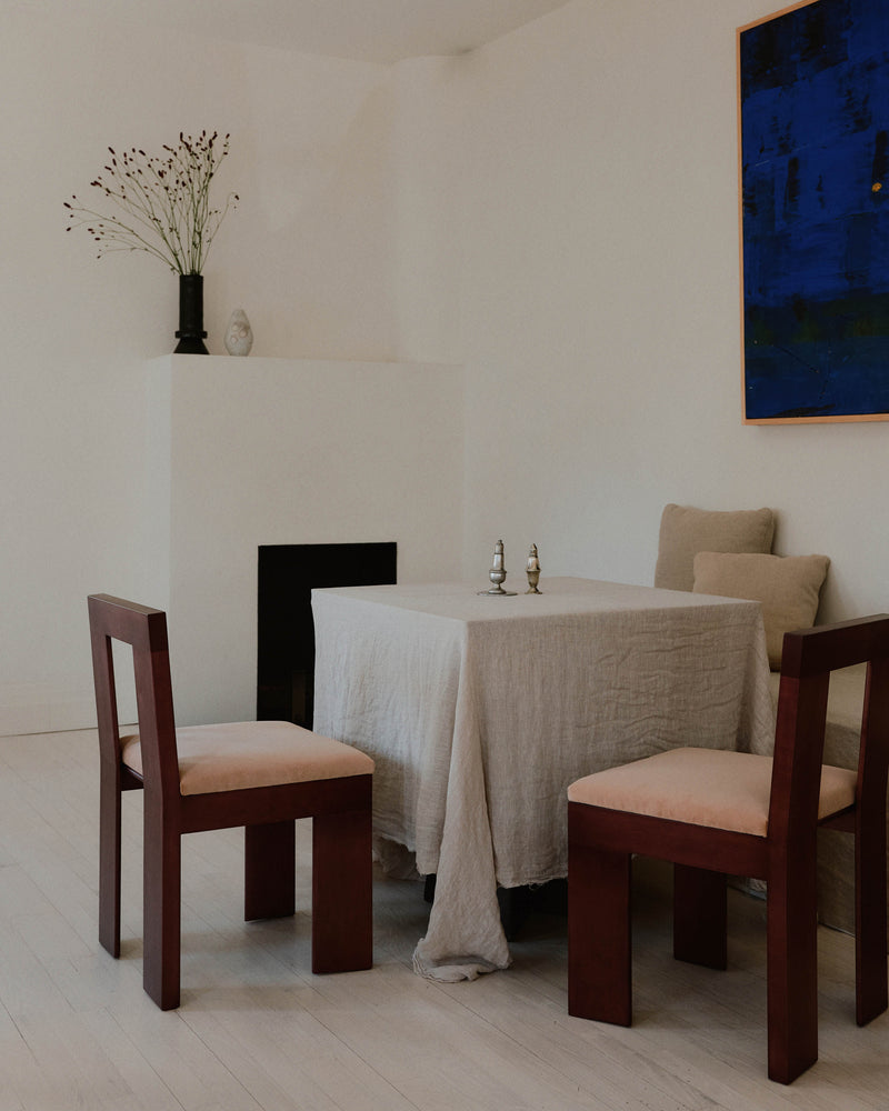 Minimalist dining room featuring contemporary furniture, including a wooden table with a light-colored tablecloth and four Raja Chairs from Juntos Projects, each with light upholstered seats. A dark vase with branches rests on a white built-in shelf, while an abstract blue painting hangs on the wall.