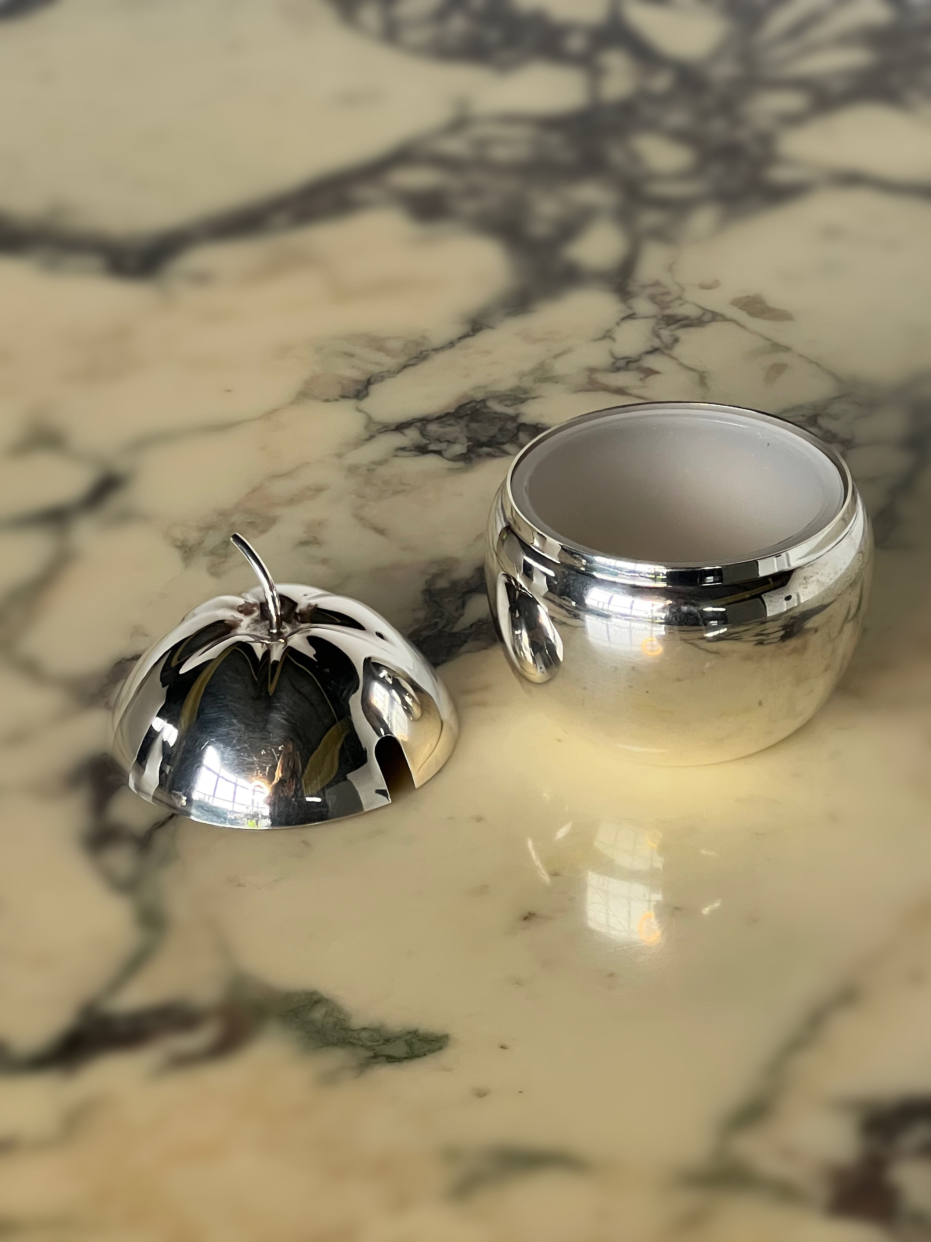 The Silver Plated Apple Jam Pot by Porter Studio, with its lid removed, reflects the intricate natural patterns of the marble surface it rests on.