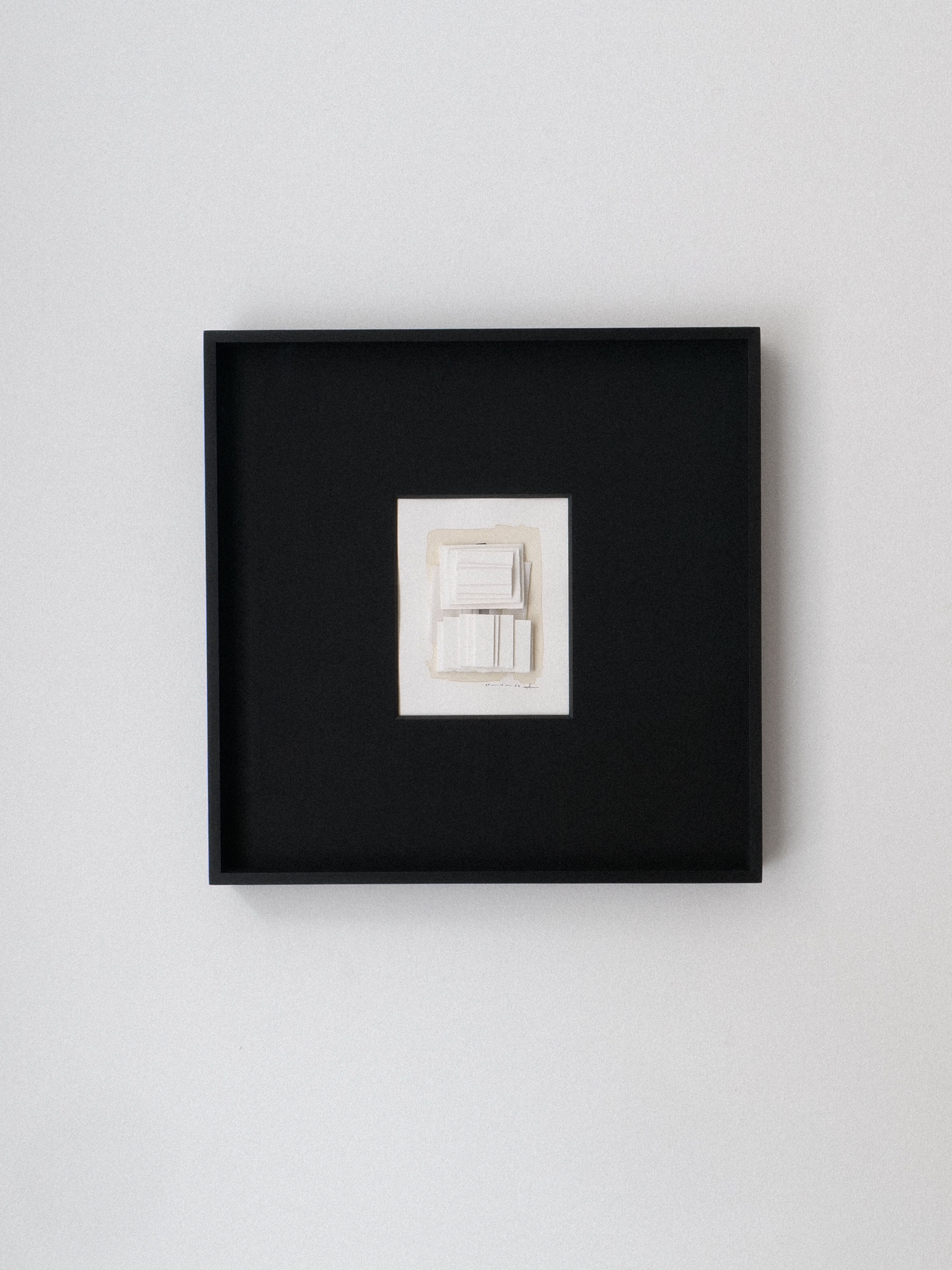 The minimalist black frame of Collage No.03 Rasmus Wingårdh by Collection apart hangs on a white wall, containing a small abstract artwork. This mixed media piece features layered white and gray geometric shapes, offering a subtle three-dimensional effect.