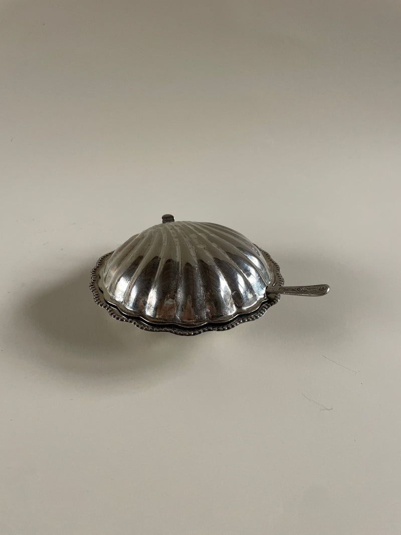 A Vintage Plated Shell Butter Dish by Maud Vaughan, featuring a silver shell shape with a small handle, gently rests on a smooth, neutral surface. This exquisite piece, resembling a clam shell with its textured, ribbed design, pairs beautifully with a vintage butter knife for an added touch of charm.