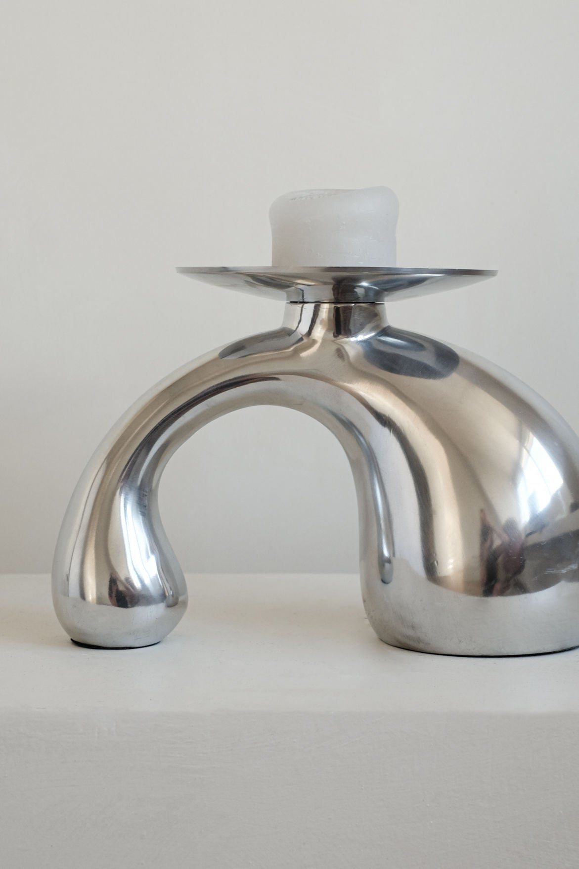 The septembre studios Bombastic ‘Drop’ Candle Holder features a curved, arch-like design in polished metal, holding a white candle—an elegant piece to enhance any modern interior.