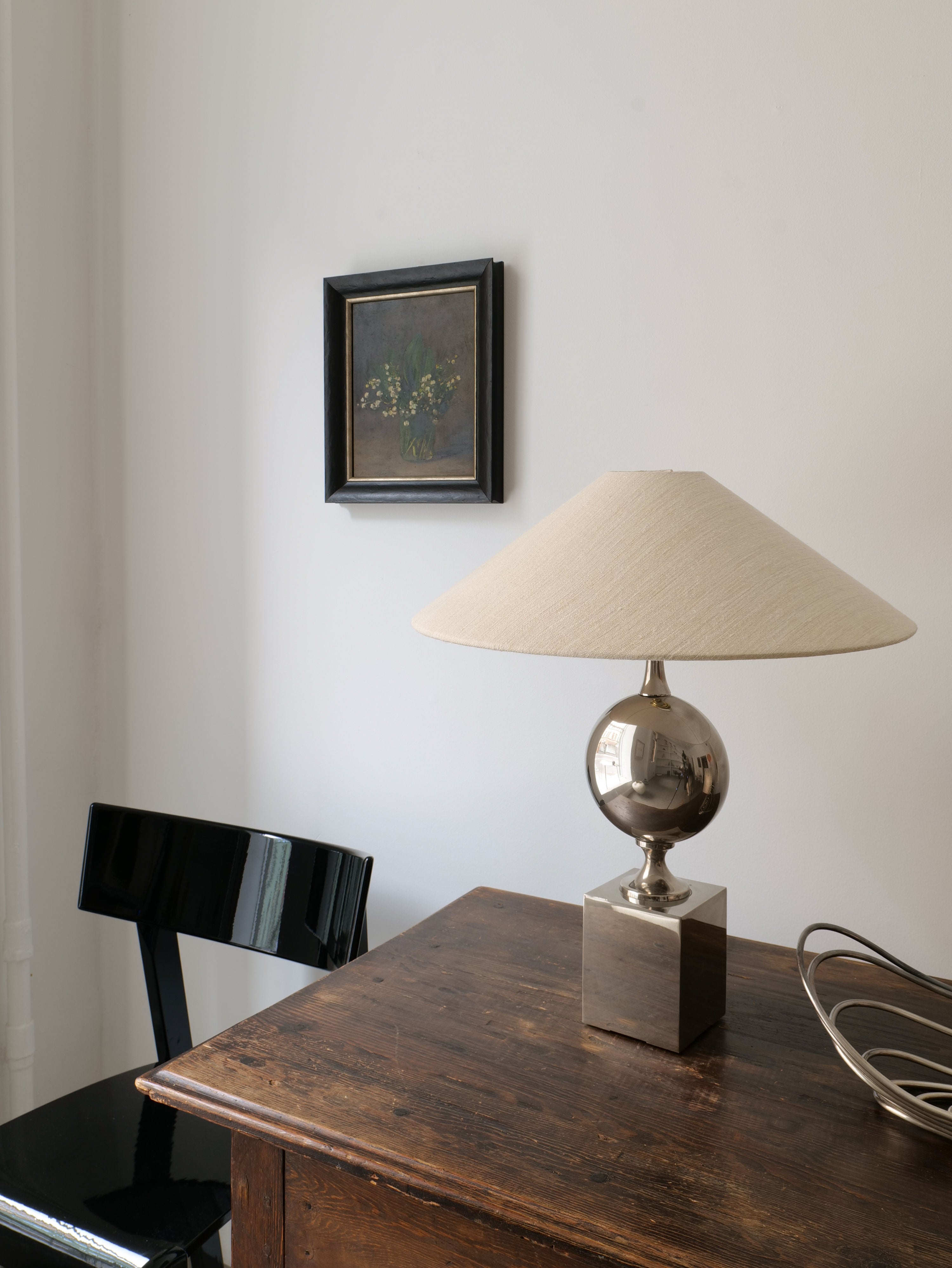 A minimalist room featuring a wooden desk with a modern silver lamp on it. A black chair stands nearby, and a framed Lily of the Valley Still Life 1961 by Collection apart hangs on the white wall above the desk. The scene is simple and tidy, with a blend of contemporary and classic elements.