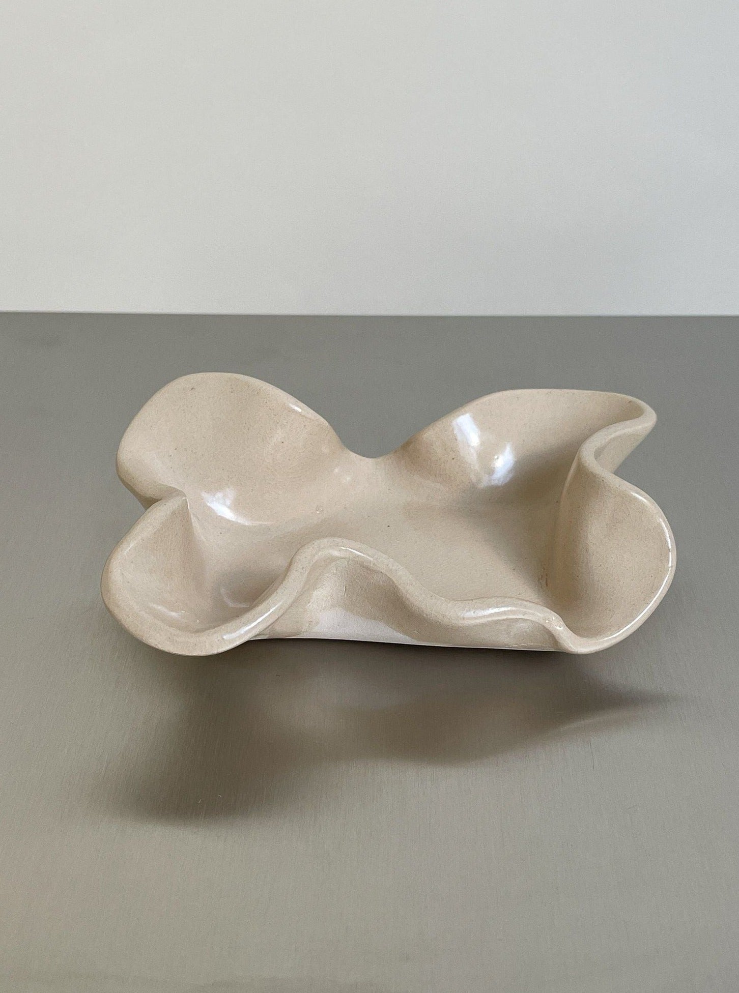 The Soapholder in stoneware by 2222Studio features a beige, wavy-edged, handmade design with an organic shape, shown on a smooth gray surface—an elegant addition to your bathroom decor.